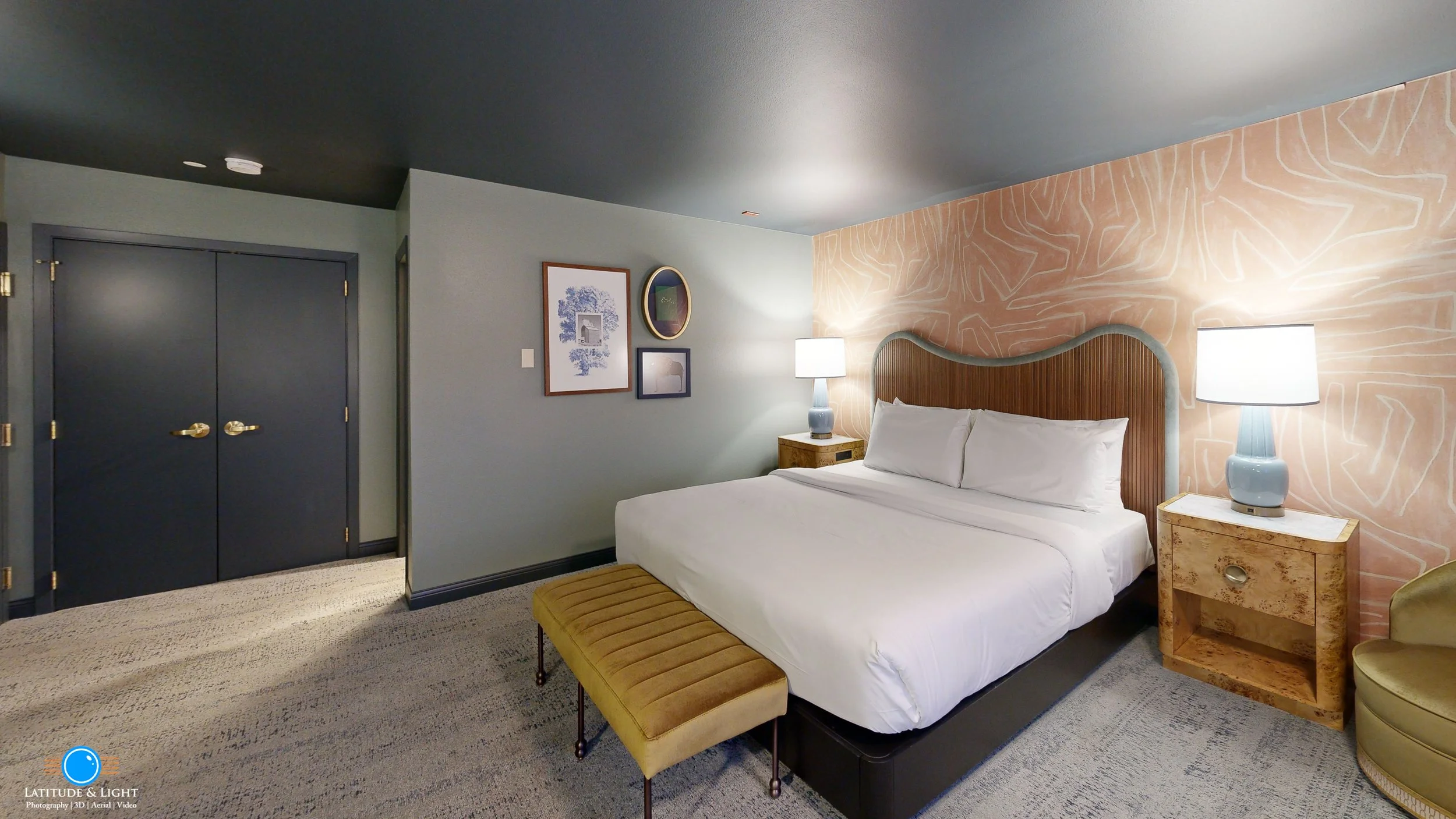 A bedroom with a large bed, two bedside tables with lamps, a green bench at the foot of the bed, and decorative artwork on the wall; twin dark doors are visible on the left.