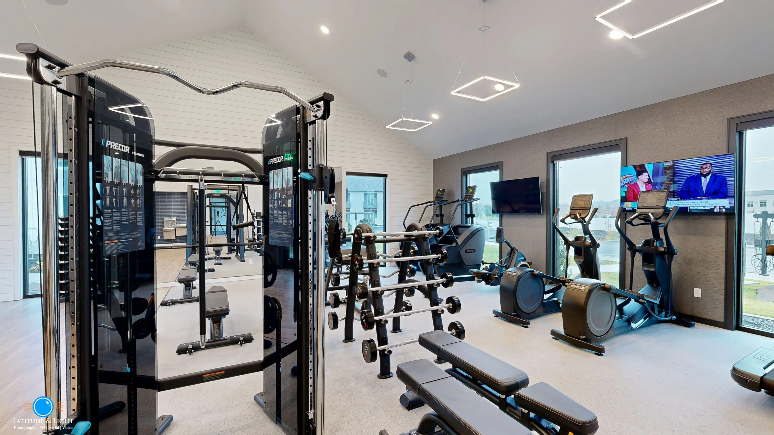 Modern gym at a Liberty Lake apartment complex with cardio and strength training equipment, including treadmills, elliptical machines, and weight racks, with large windows and two TV screens.