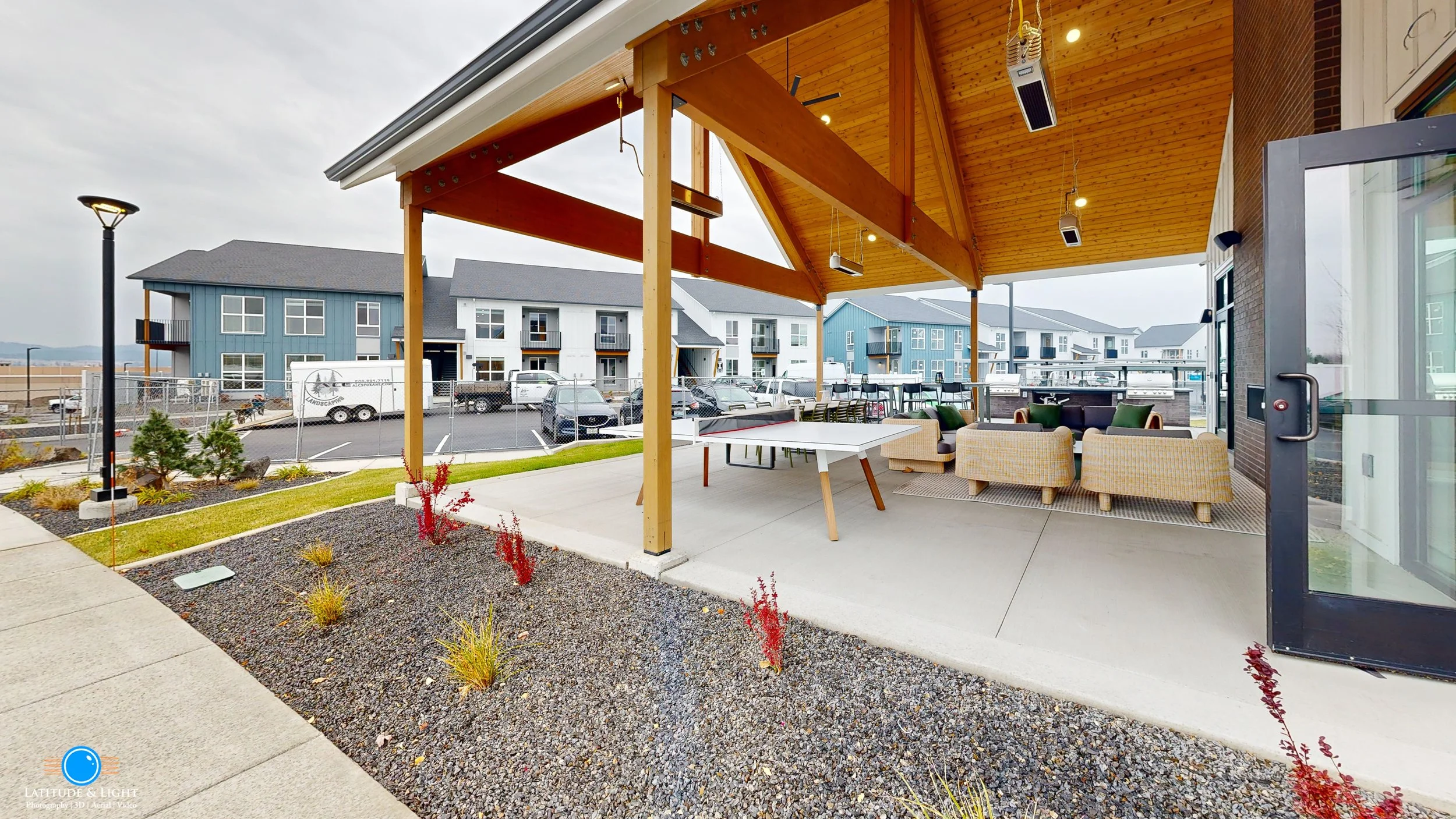 Outdoor patio at Spokane area apartment complex with seating area under a wooden pergola, with a concrete floor, containing sofas, chairs, and tables, overlooking a parking lot and apartment buildings, with garden beds and a nearby sidewalk.