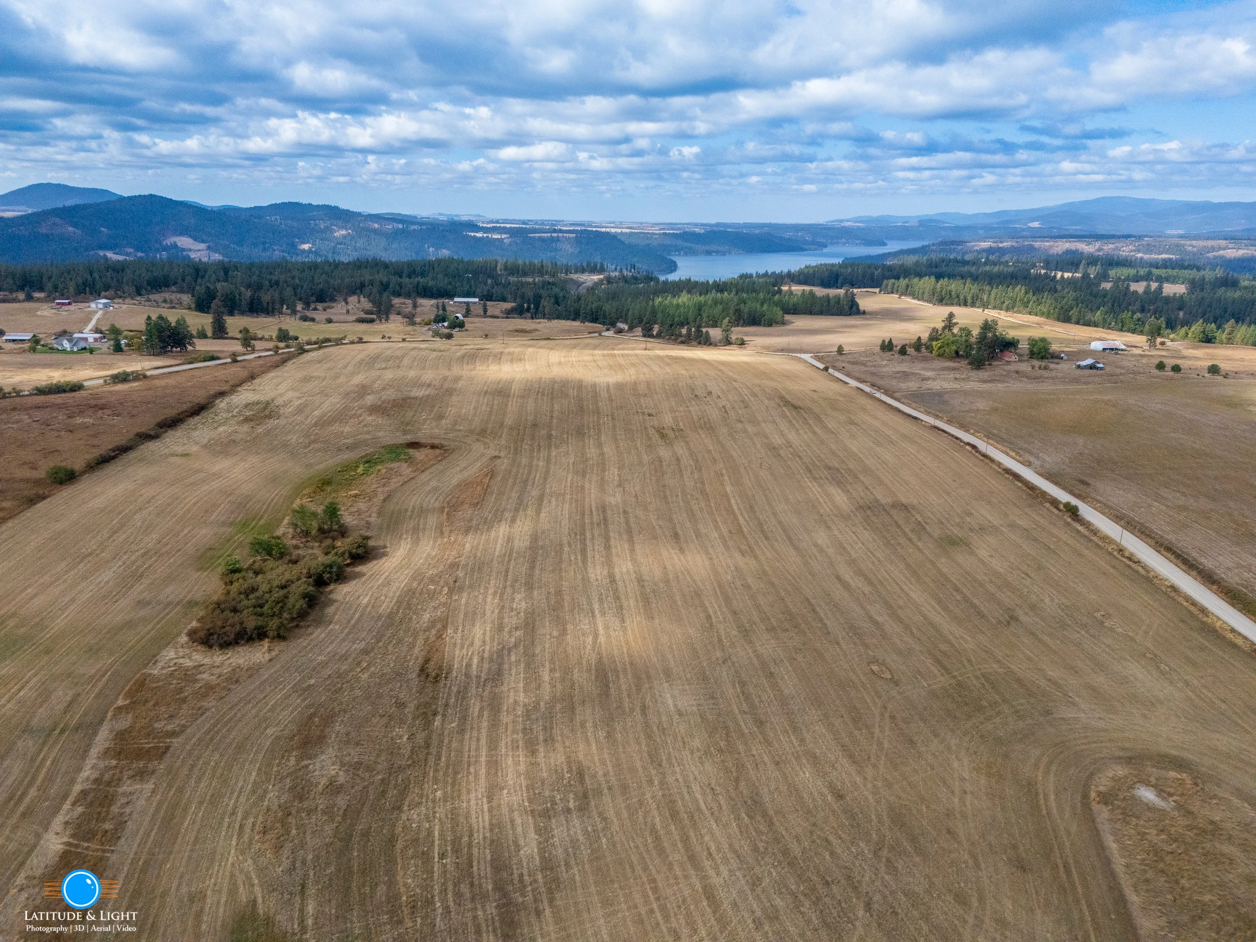 Aerial view of rural farmland with fields, trees, houses, a road, and a lake in the background under a partly cloudy sky.