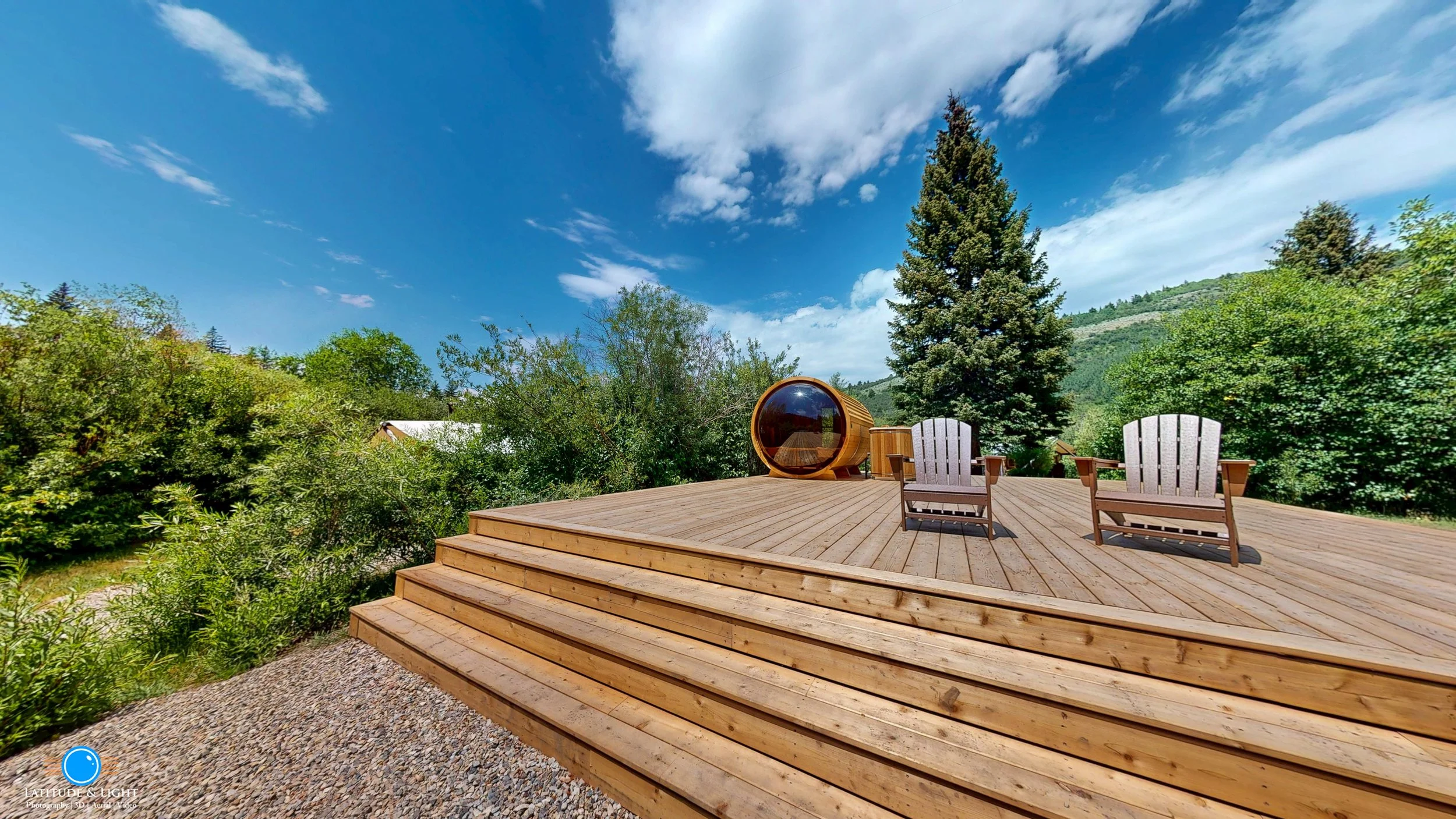 A wooden deck at a Victor, Idaho resort with two Adirondack chairs and a small side table, set against a backdrop of green trees, a hill, and a blue sky with white clouds, with an outdoor pod in the background.