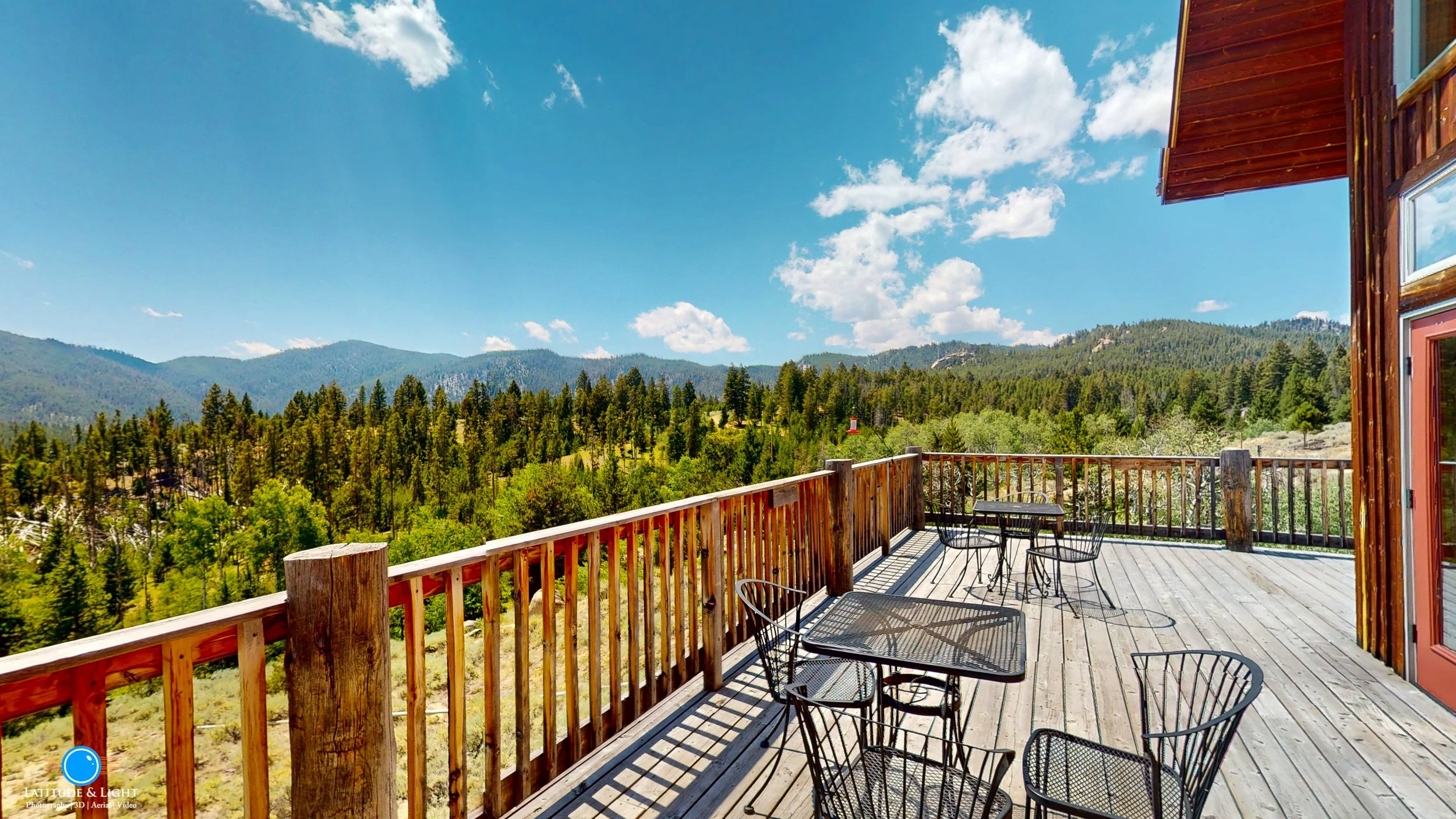 Outdoor wooden deck with metal tables and chairs overlooking a forested mountain landscape under a partly cloudy sky.