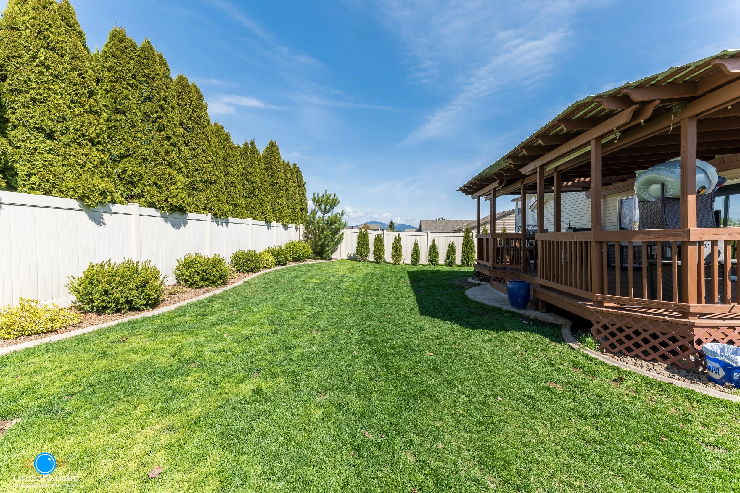 Post Falls, Idaho Backyard with green grass, landscaped bushes, and tall trees behind a white fence. There is a covered wooden deck attached to a house with outdoor furniture and a blue planter.