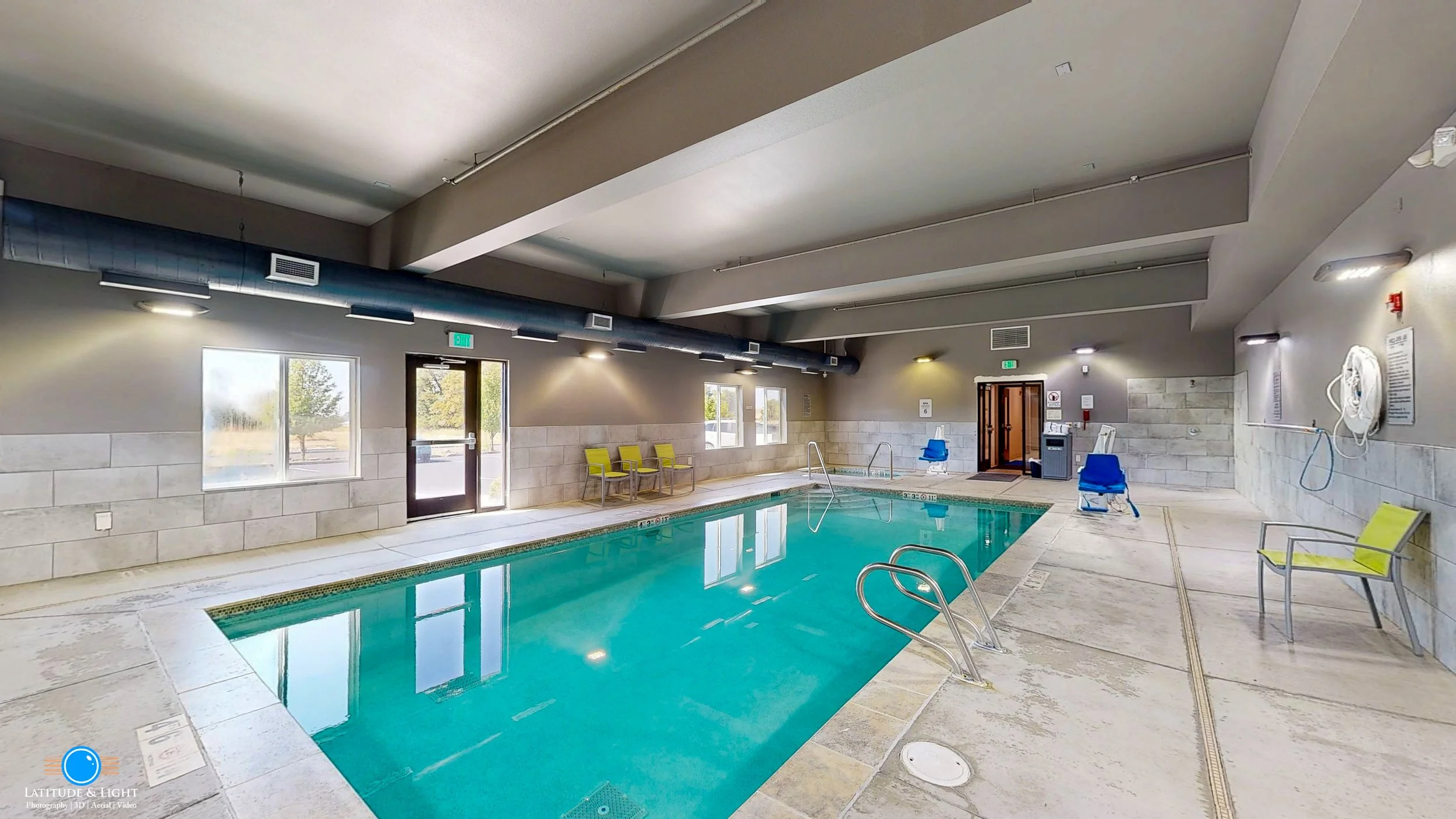 Indoor swimming pool at a Walla Walla hotel with metal handrails, yellow and blue chairs, and large windows letting in natural light.