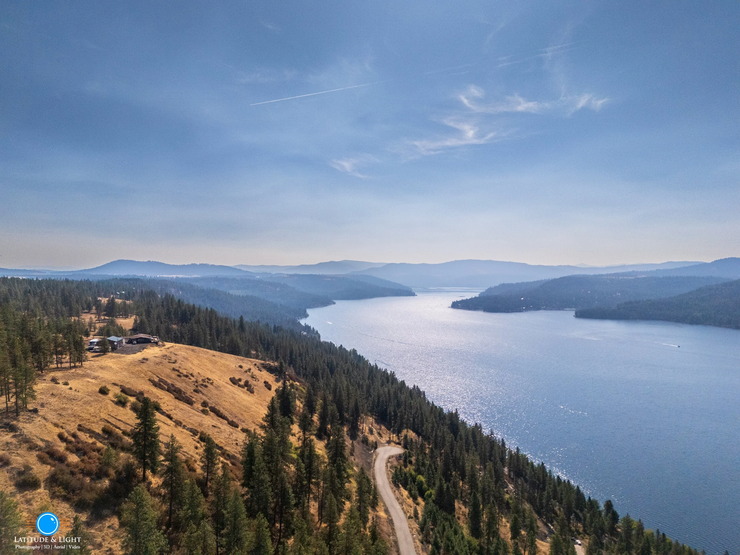 A scenic view of a lake with surrounding forested hills and a winding road near the shore on a clear day.