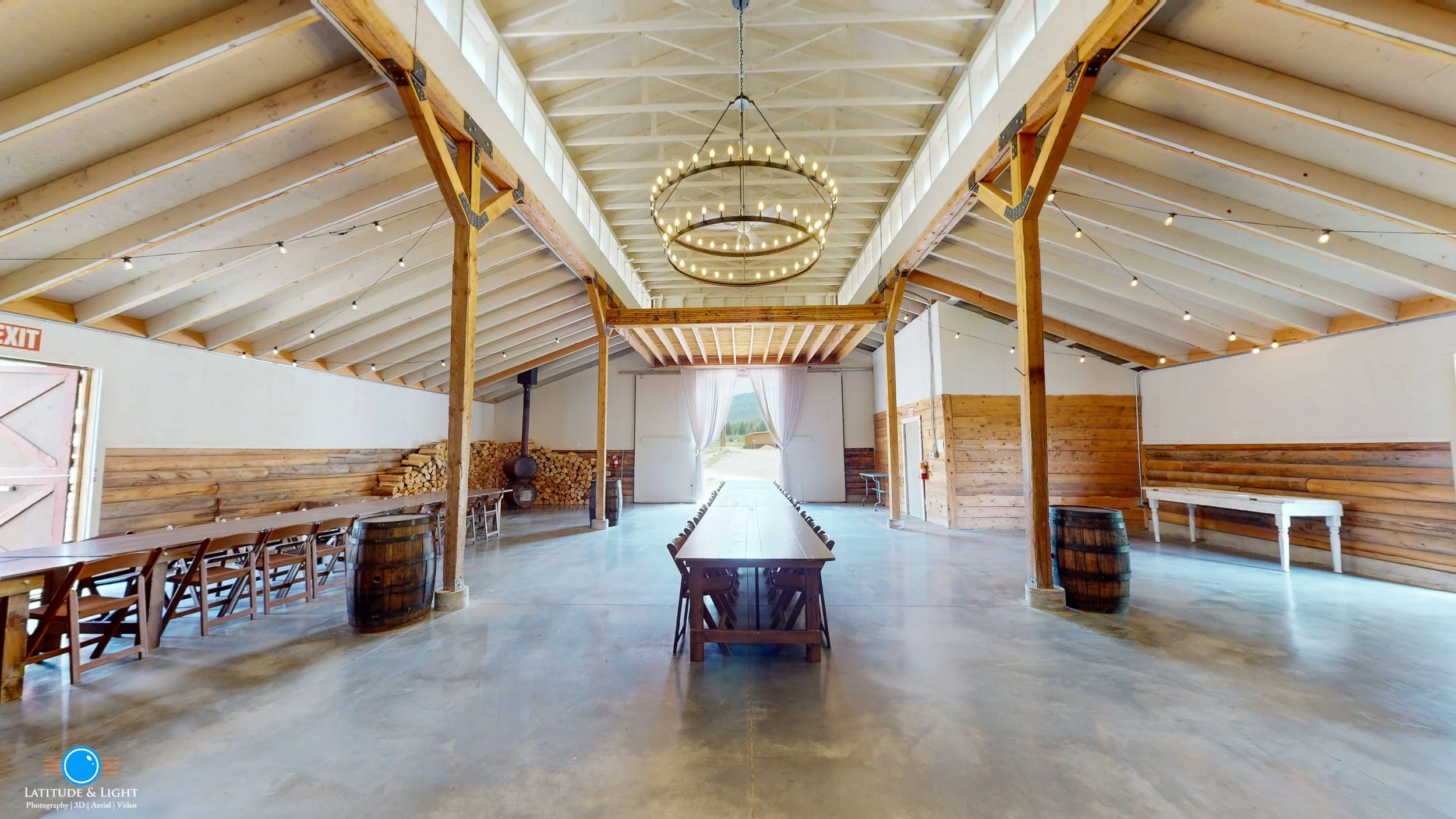 Interior of a rustic event barn in Montana with high vaulted ceilings, string lights, a central long wooden table with chairs, and a decorative circular chandelier, wood-burning stove, wood pile, and large open doors revealing an outdoor view.