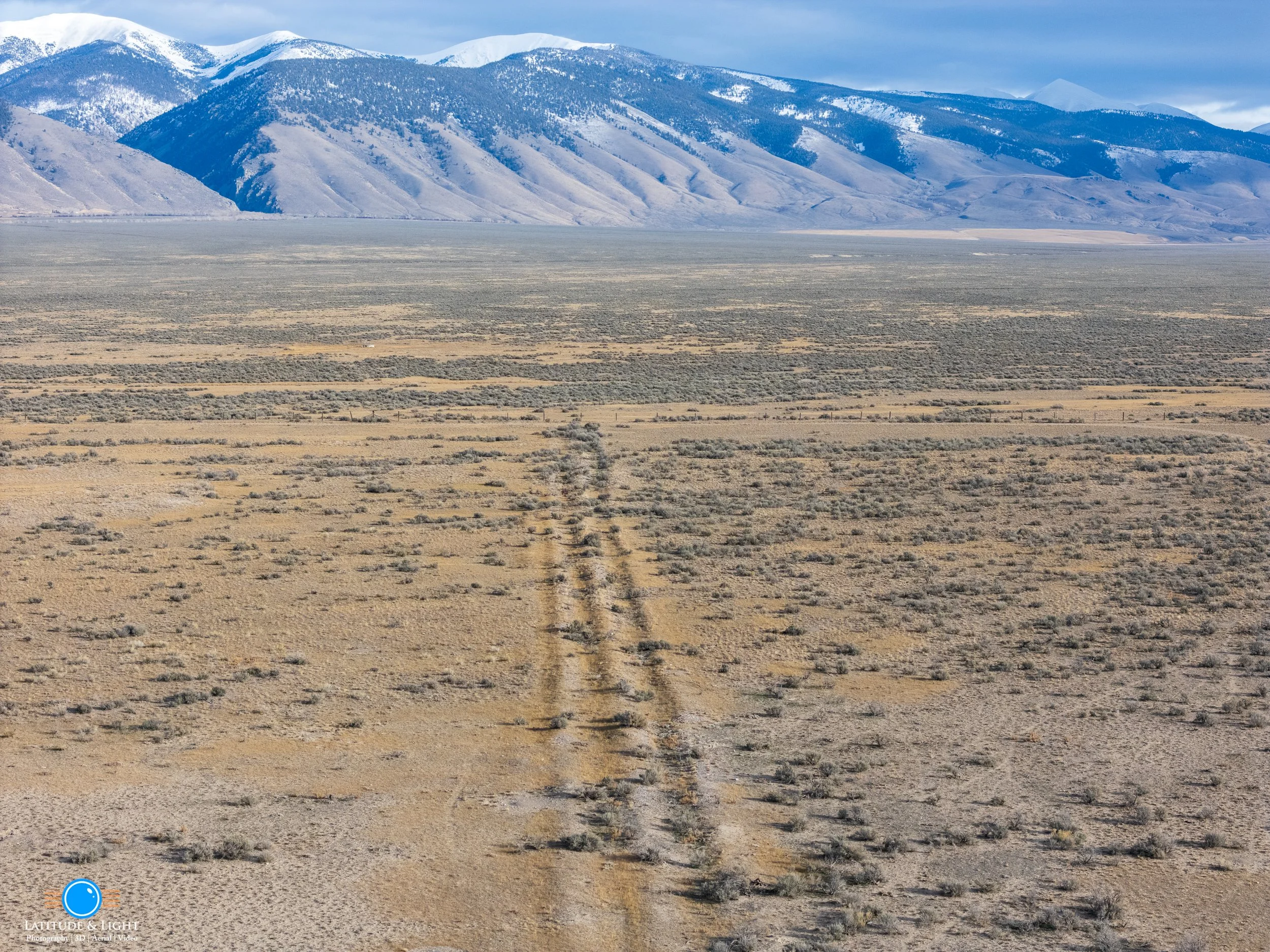 A vast desert landscape with sparse vegetation and a dirt trail leading towards distant snow-capped mountains under a cloudy sky.