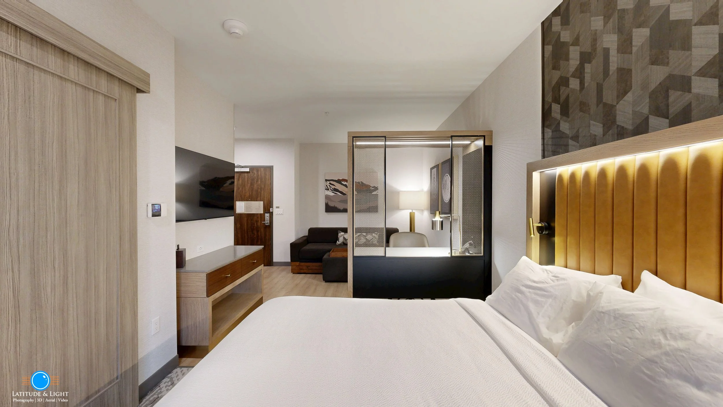 Modern hotel room in Sandpoint, Idaho with a king-sized bed, a textured headboard, and a partial view of a sitting area with a sofa, TV, and artwork in the background.