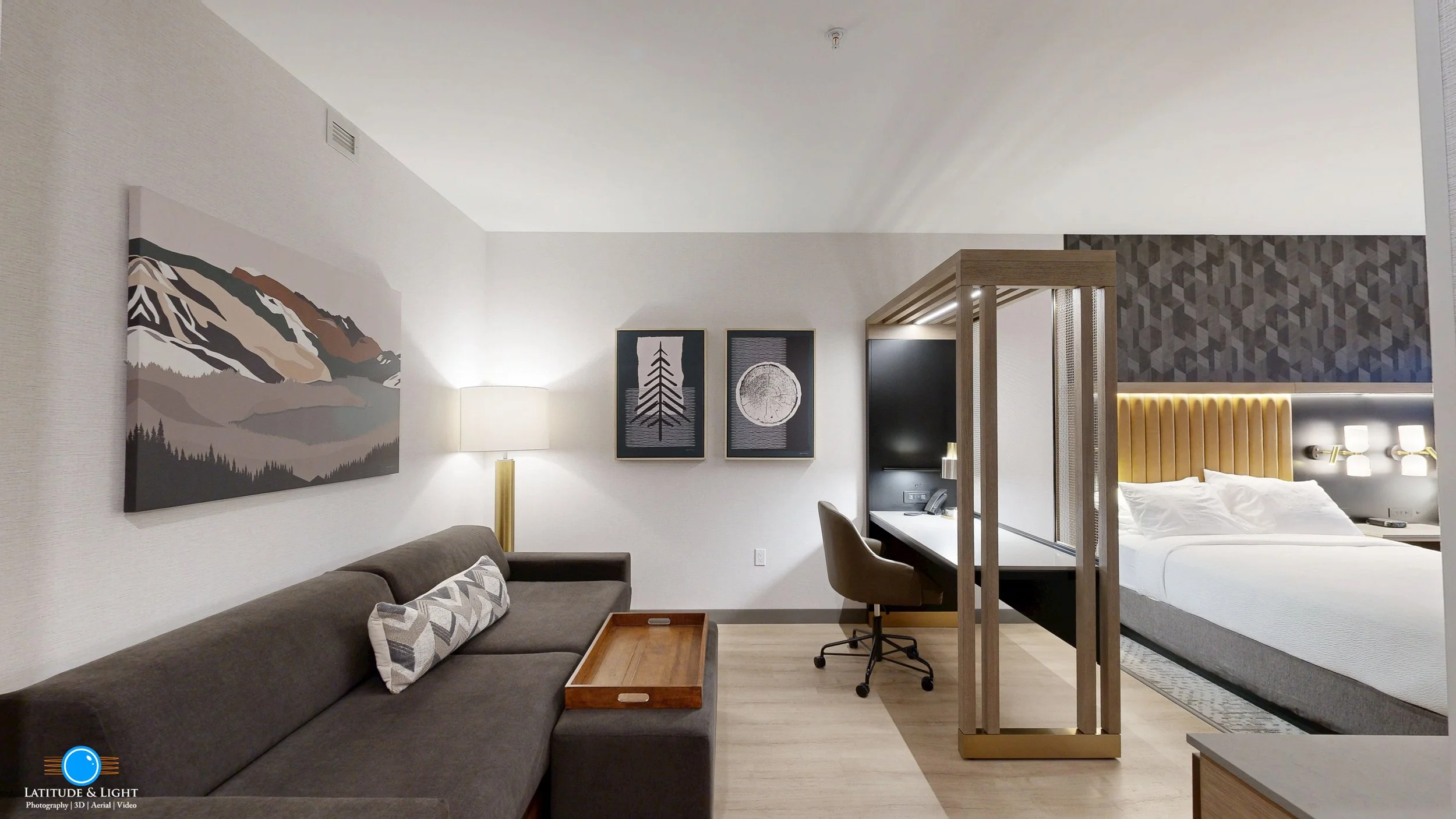 A modern hotel room with a grey sofa, a white lamp, and wall art depicting mountains, trees, and a moon. There is a work desk with a chair, and a sleeping area with a bed and yellow tufted headboard. The room features neutral colors and contemporary 