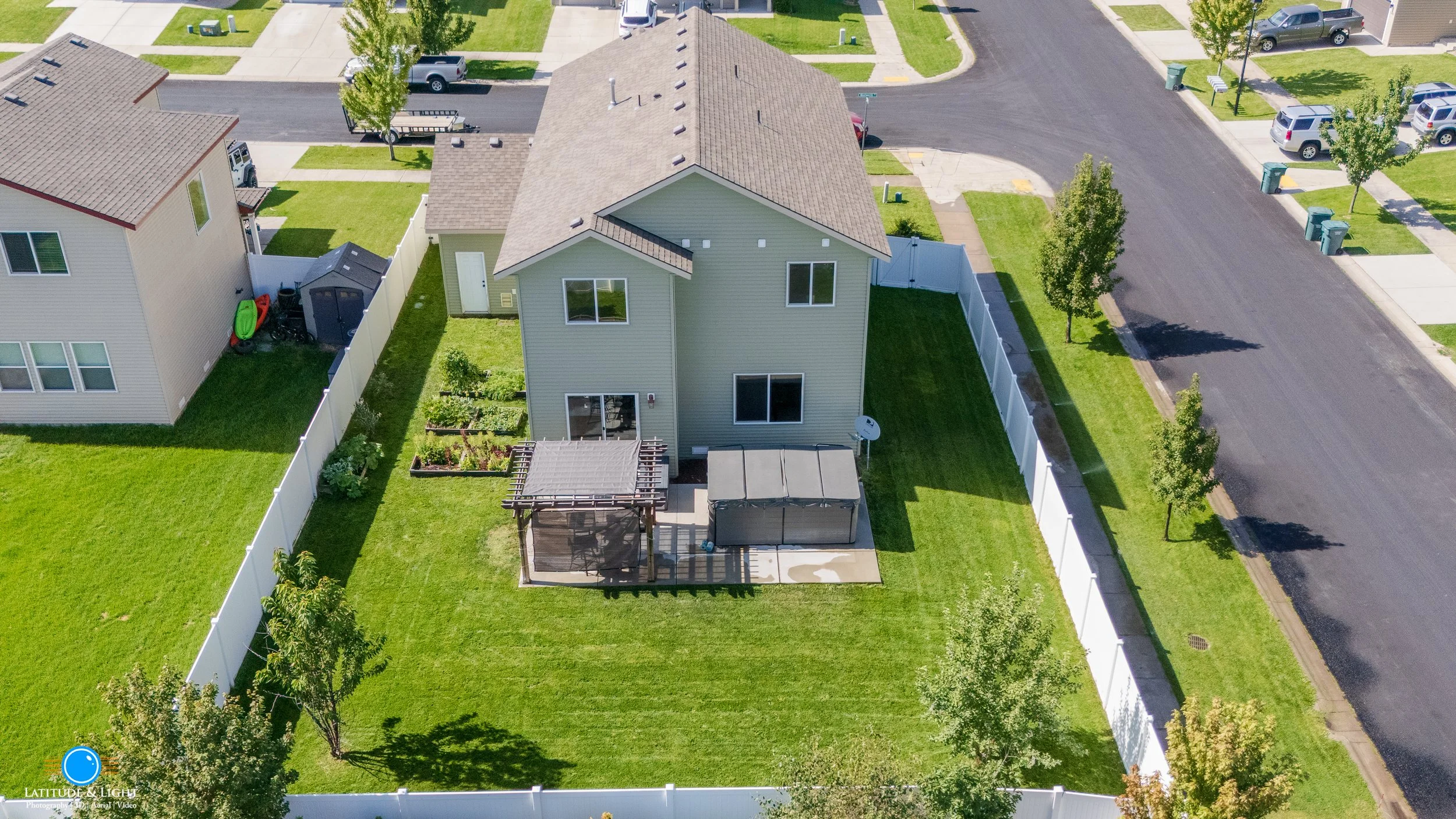 Aerial view of a Post Falls, Idaho backyard with green grass, a pergola, and a hot tub, enclosed by a white fence, in a suburban neighborhood.