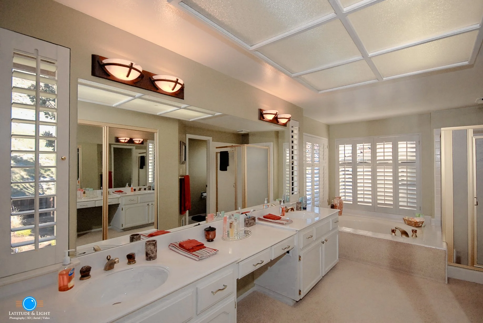 Bright Coeur D'Alene bathroom with large vanity mirror, white cabinets, two sinks, and double light fixtures. Multiple windows with white shutters, a bathtub, and a separate shower stall are visible.