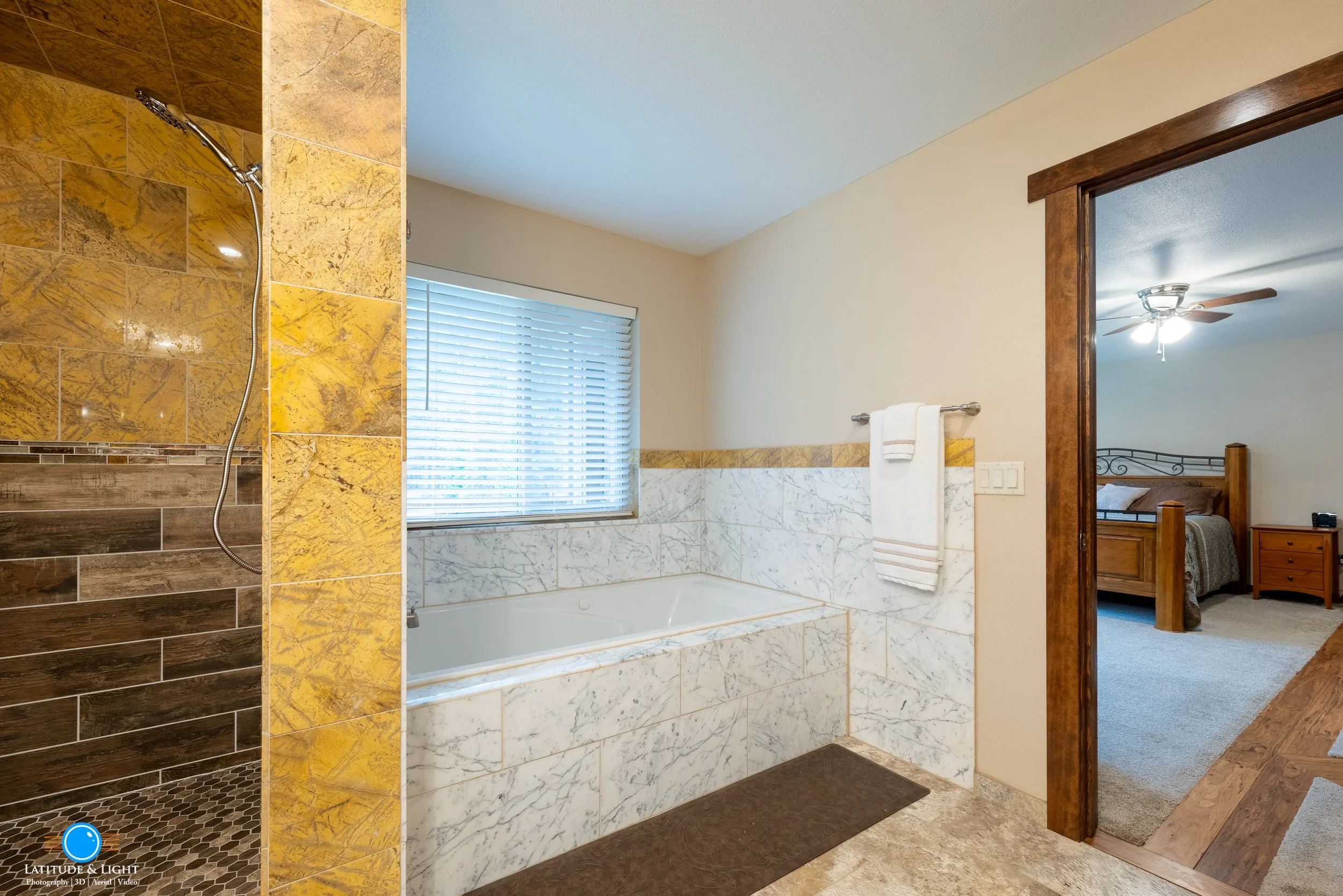Bathroom with bathtub and shower area. The shower has brown and yellow tiles, and the bathroom has marble tiles around the bathtub. A window with blinds is above the bathtub, and a white towel hangs on a metal rack on the wall. The bathroom opens int