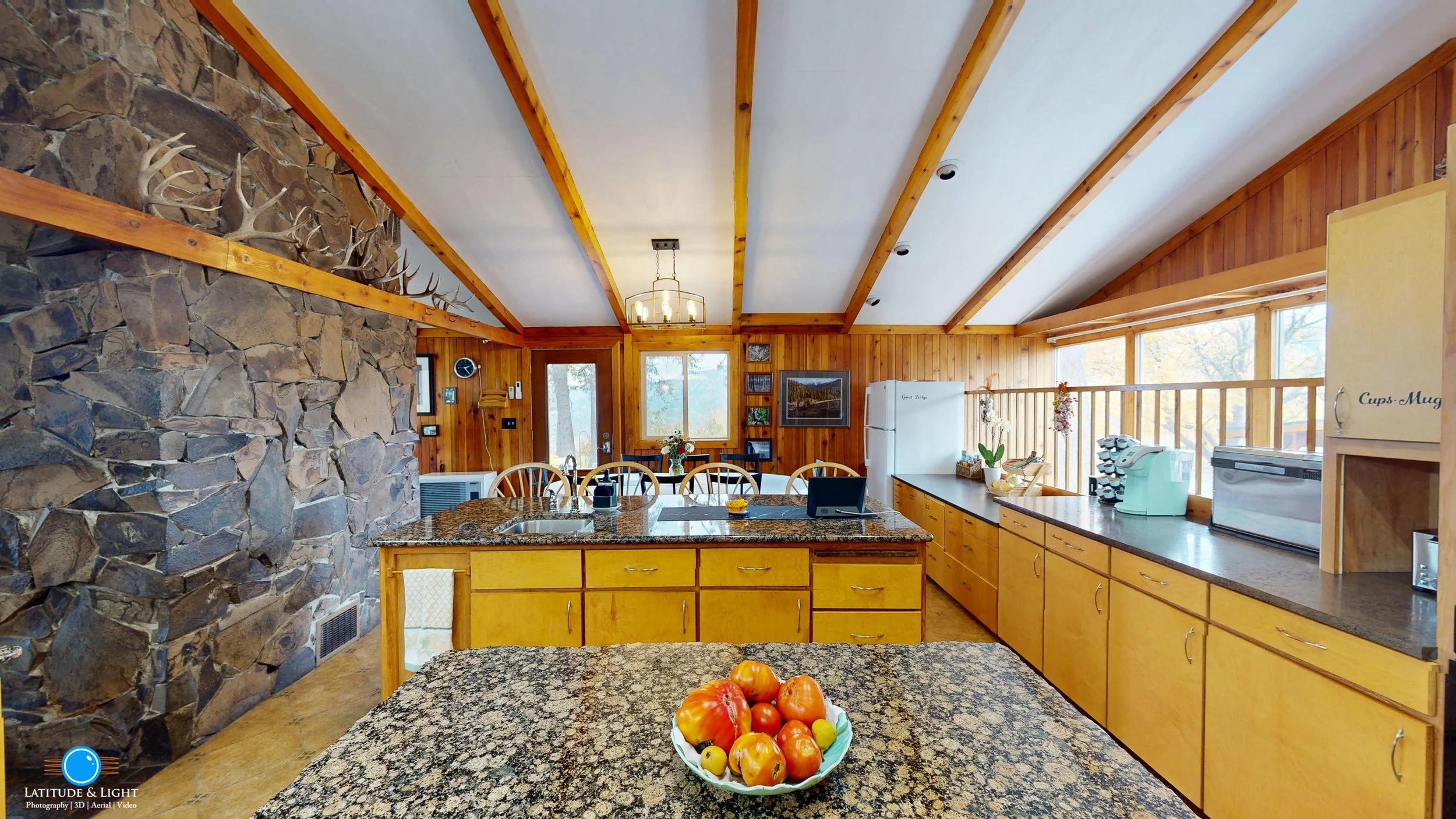 A rustic kitchen in a rural Montana wedding ranch with wooden walls, ceiling beams, granite countertops, and a stone accent wall with antlers. There is a bowl of apples on the island, a white refrigerator, and large windows letting in natural light.