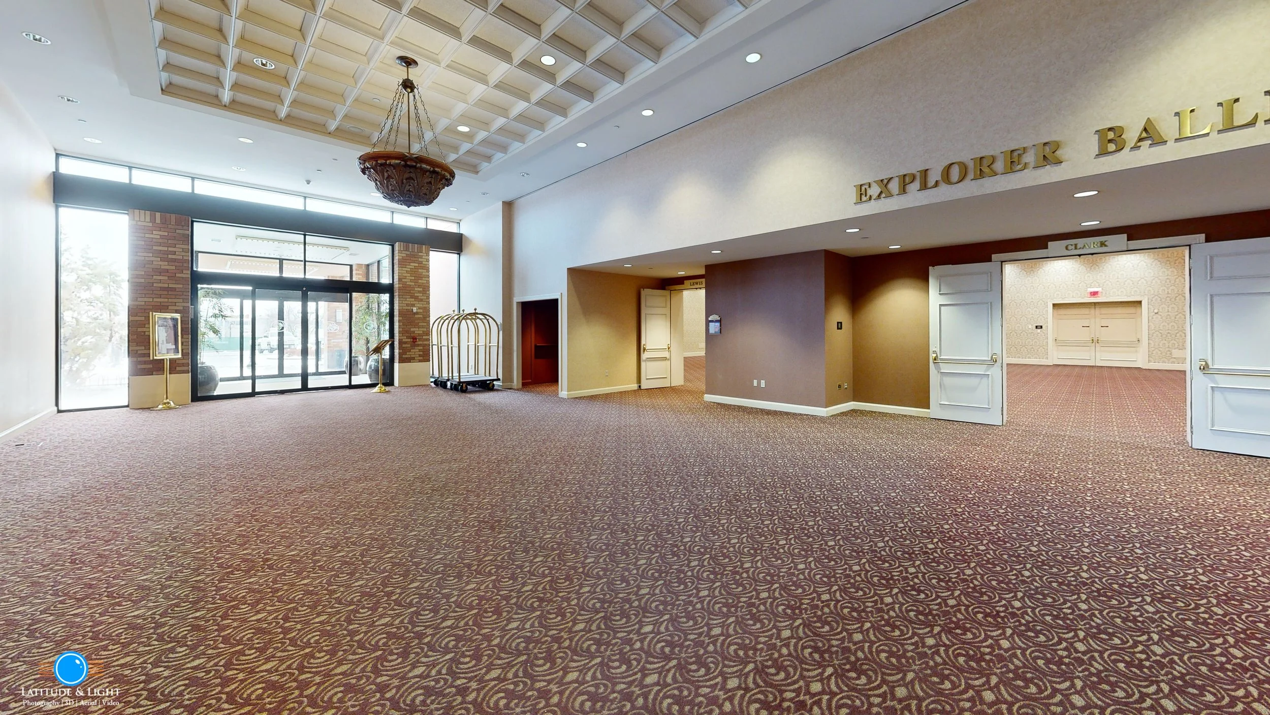 Empty hotel ballroom in Walla Walla with patterned carpet, high ceiling with decorative chandelier, front entrance with glass doors, and signs for Explorer Ballroom.