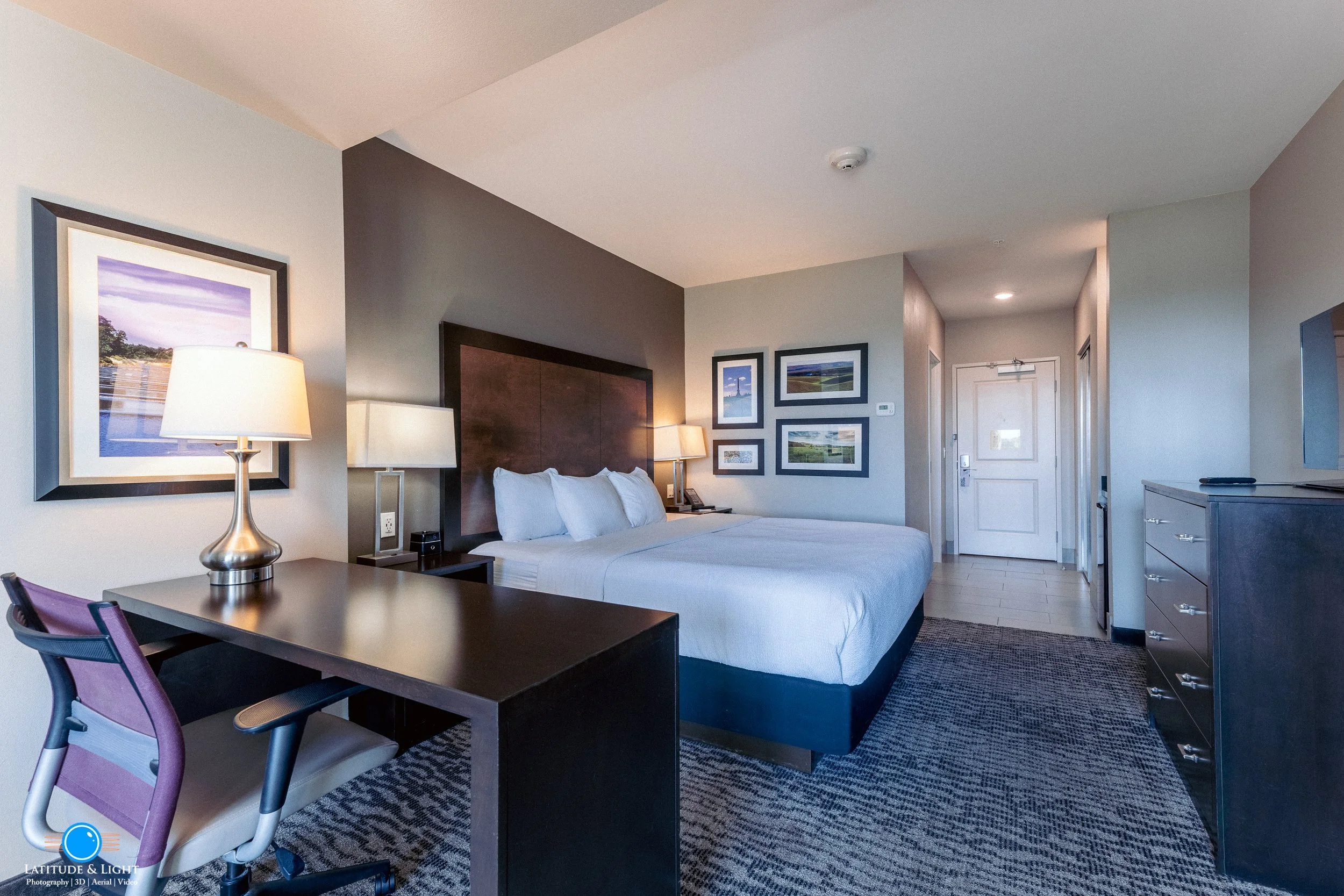 A hotel room with a large bed, nightstand, desk with a lamp and chair, framed pictures on the wall, dresser, and an entrance door.