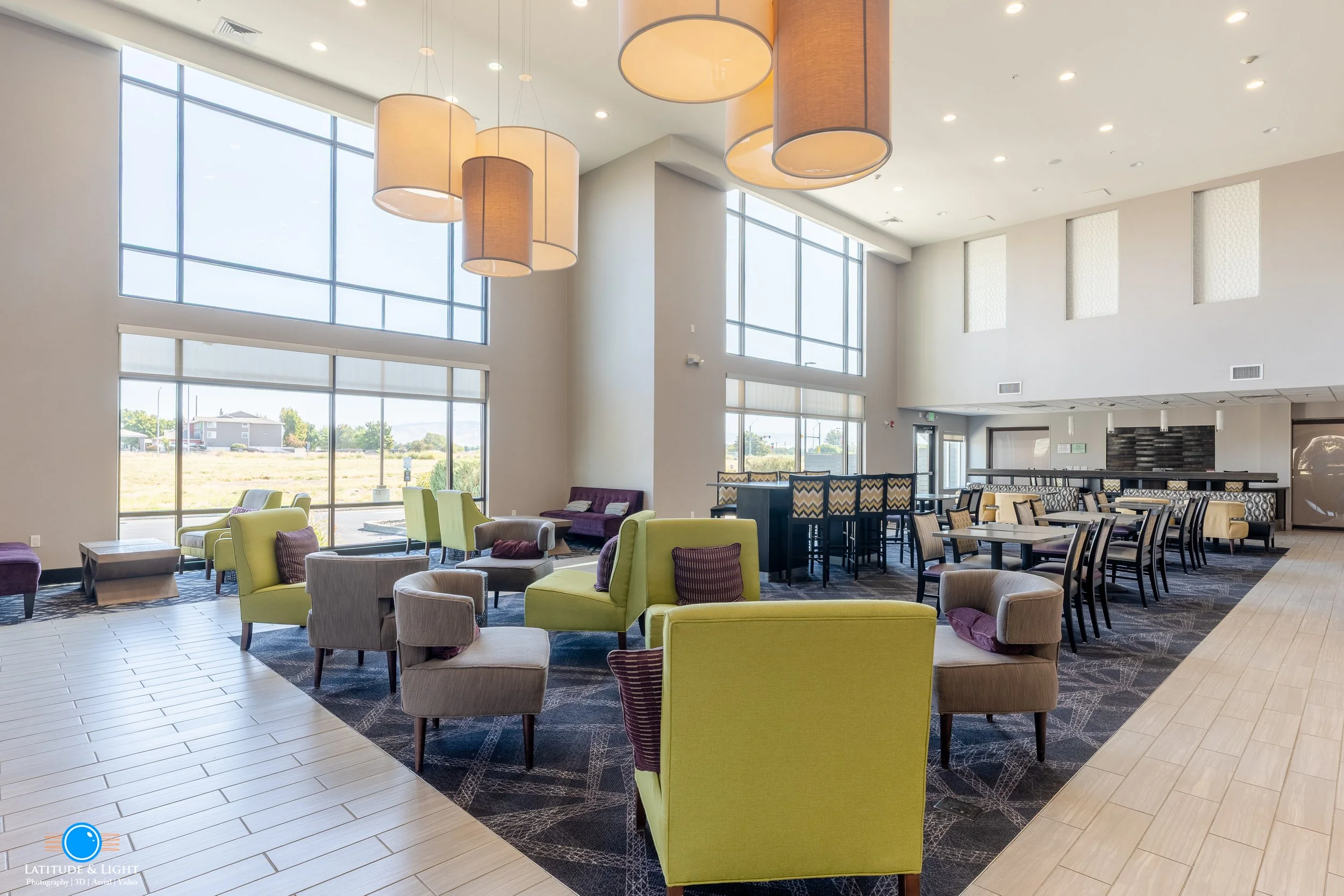LaQuinta Inn by Wyndham in Walla Walla features a modern and spacious lounge area with large floor-to-ceiling windows, contemporary chairs, sofas, and a high ceiling with hanging cylindrical pendant lights.