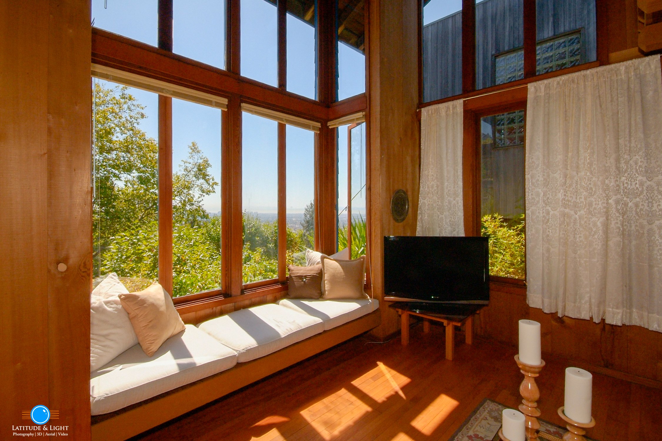 Sunlit room with large windows, a window seat with cushions, a small television on a wooden stand, and white curtains, with greenery outside