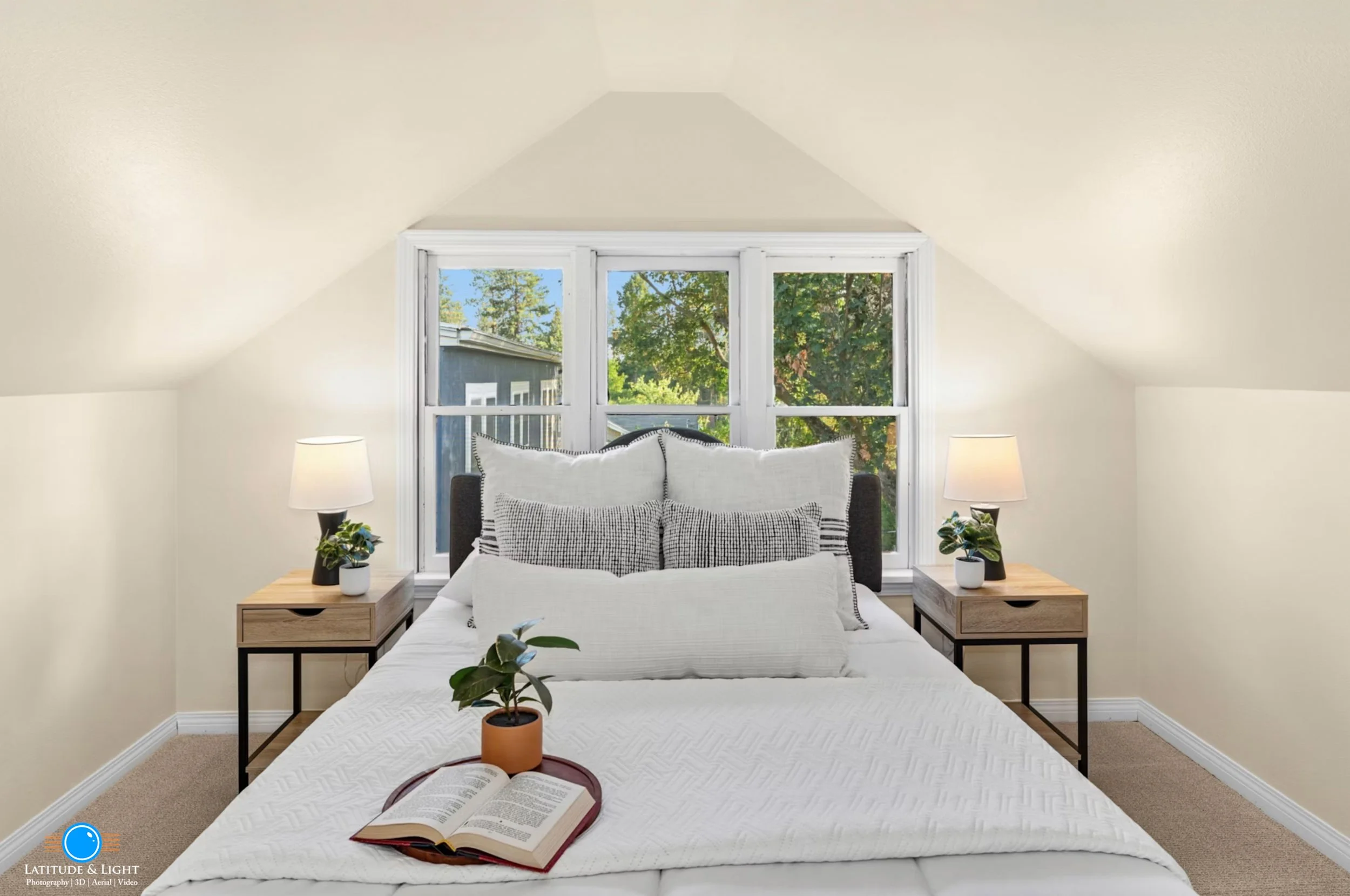 Spokane: A cozy bedroom with a cathedral ceiling, a bed with white bedding and multiple pillows, two matching nightstands with lamps, and large windows showing trees outside.