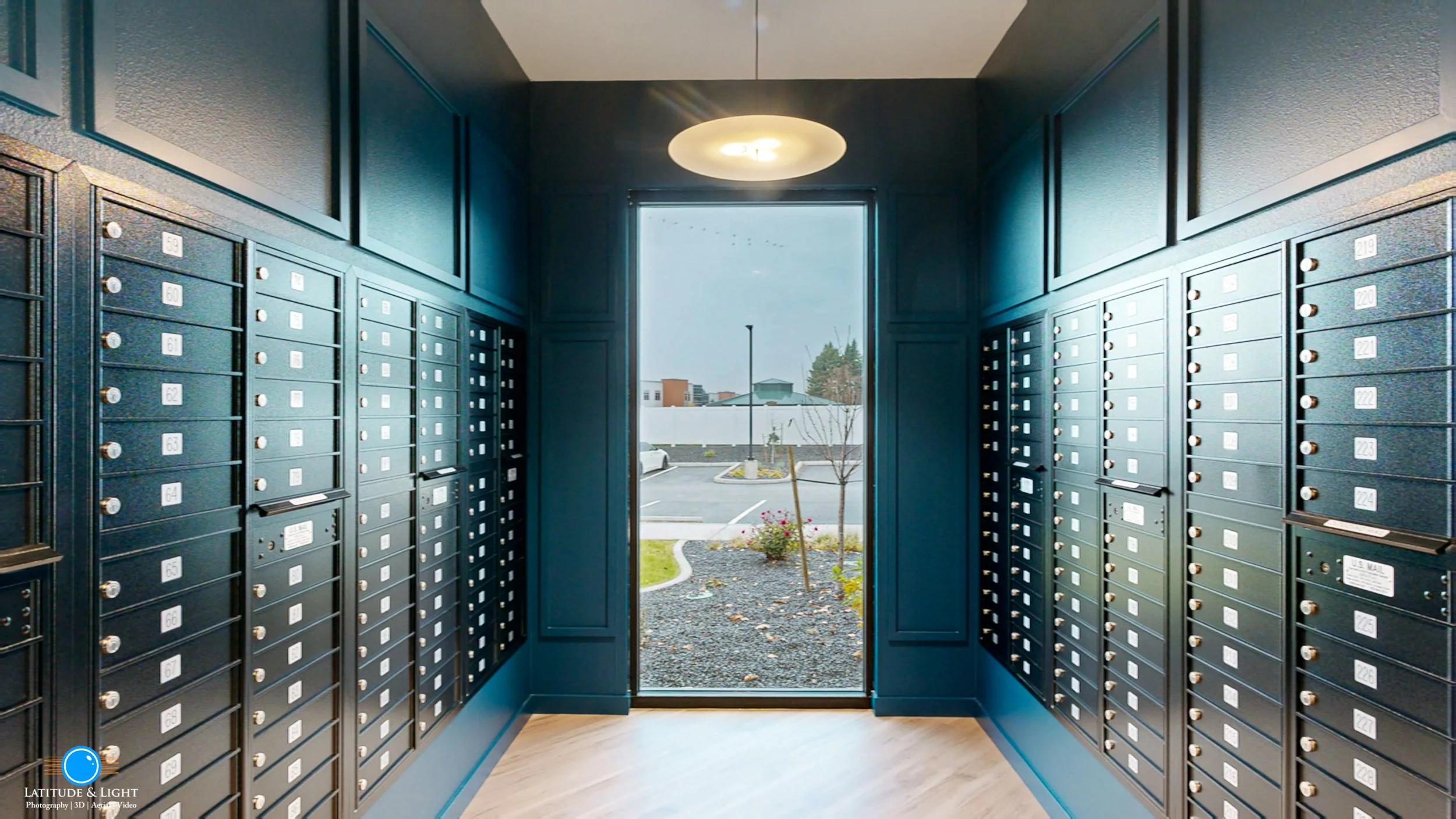 Interior of a Spokane Valley apartment mailbox area with rows of black mailboxes on both sides and a glass door leading outside, showing a parking lot and some buildings.
