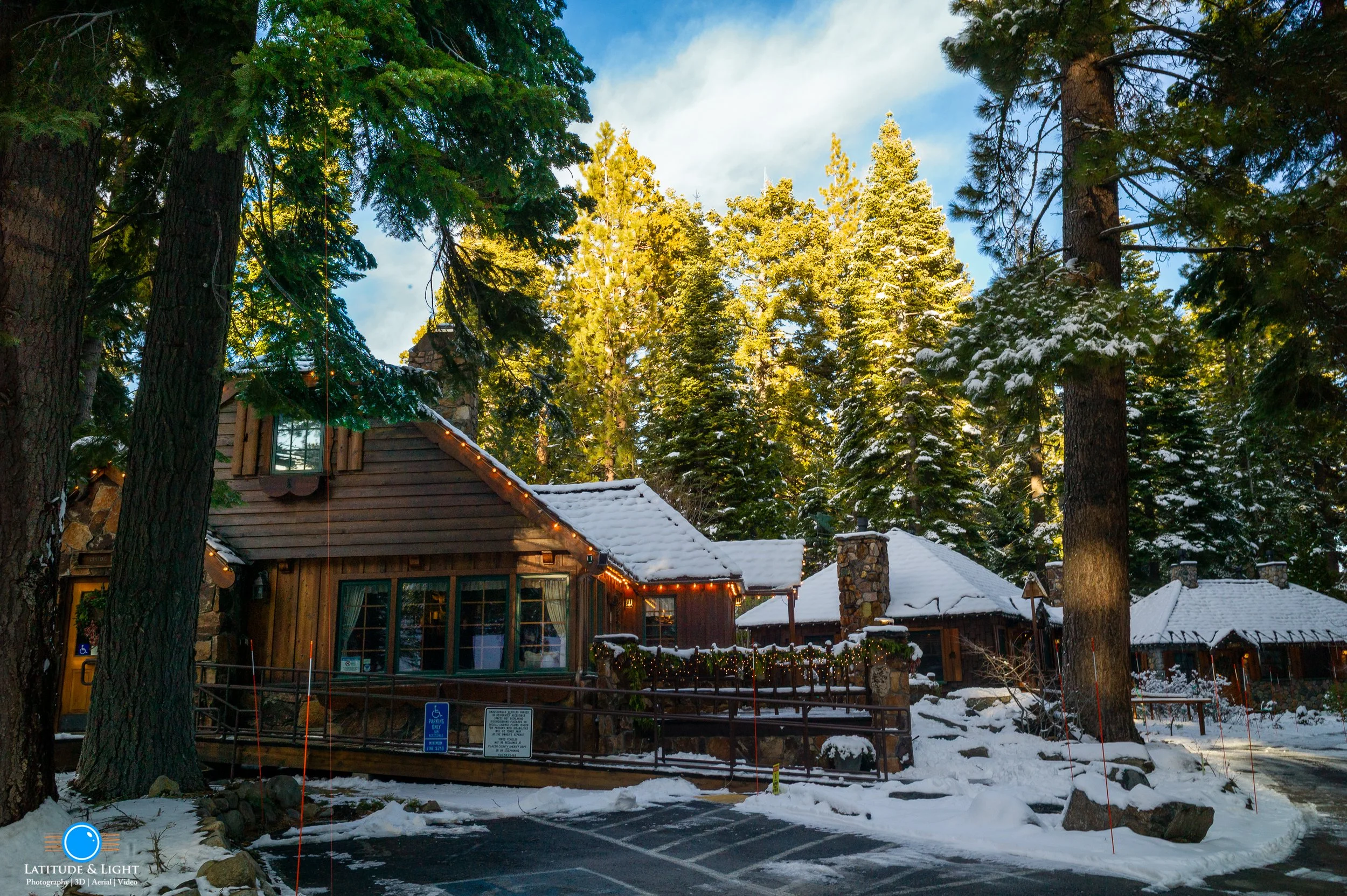 The Lodge at Tahoe City's Cottage Inn is a snow-covered log cabin surrounded by tall evergreen trees with snow on their branches, decorated with string lights, under a partly cloudy sky.