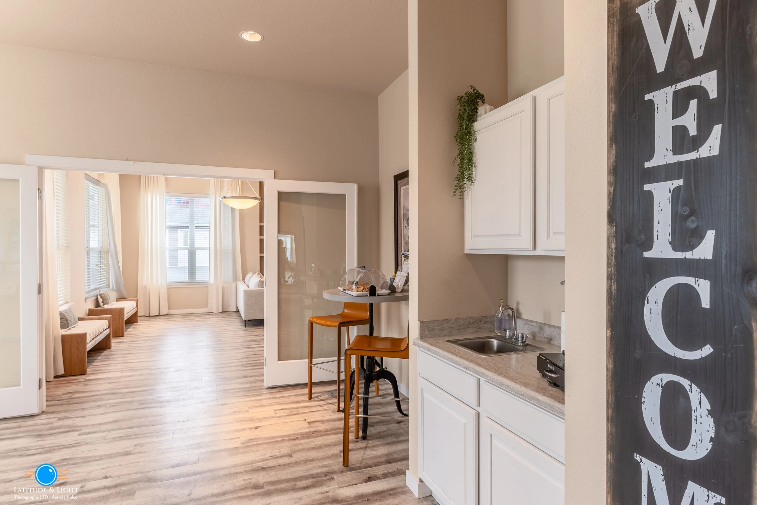 A Liberty Lake, WA apartment community center has a large living room and kitchen area with white cabinets, a small counter with two brown chairs, and a sign with the word 'WELCOME' on the right.