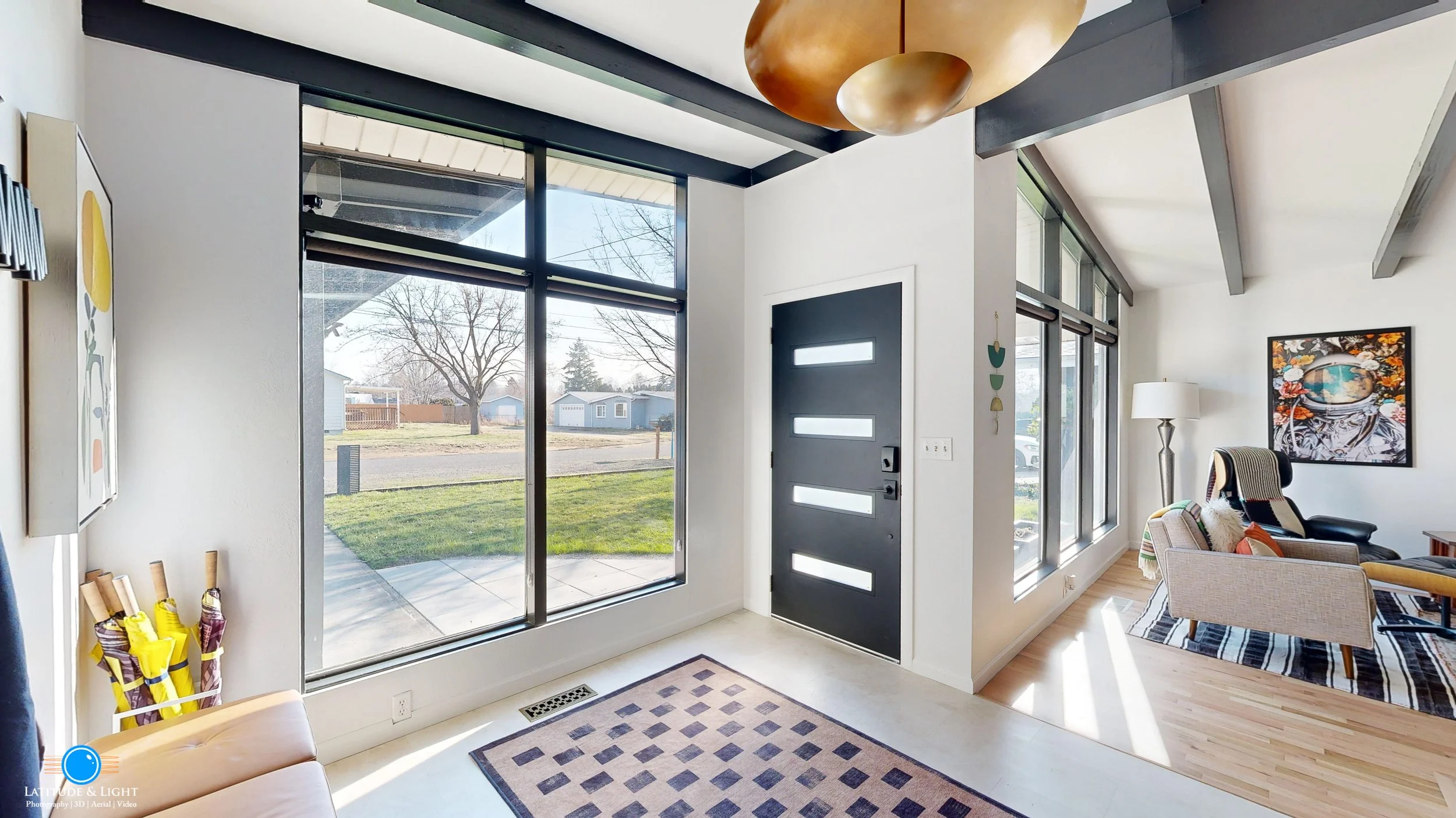 Bright front entryway with large glass windows, black front door with rectangular glass panels, a patterned rug on a white floor, and part of a living room with a black leather chair, beige armchair, artwork, and a floor lamp visible.