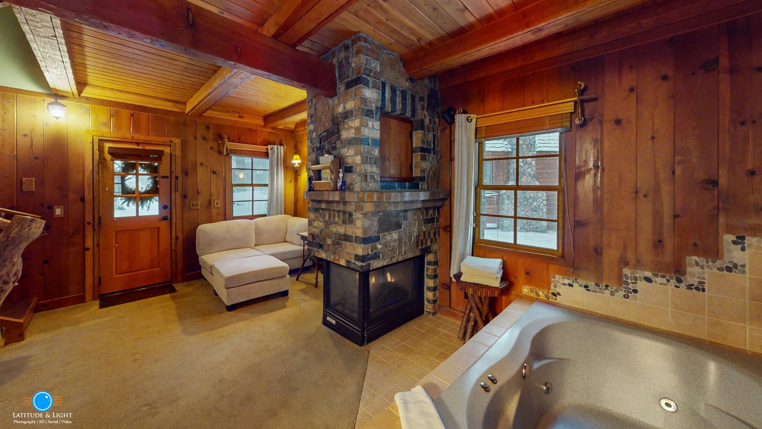 A cozy living room with wood-paneled walls, a stone fireplace, and a hot tub in the corner. There's a white sofa, windows with curtains, and a small side table with towels, all illuminated by warm lighting.