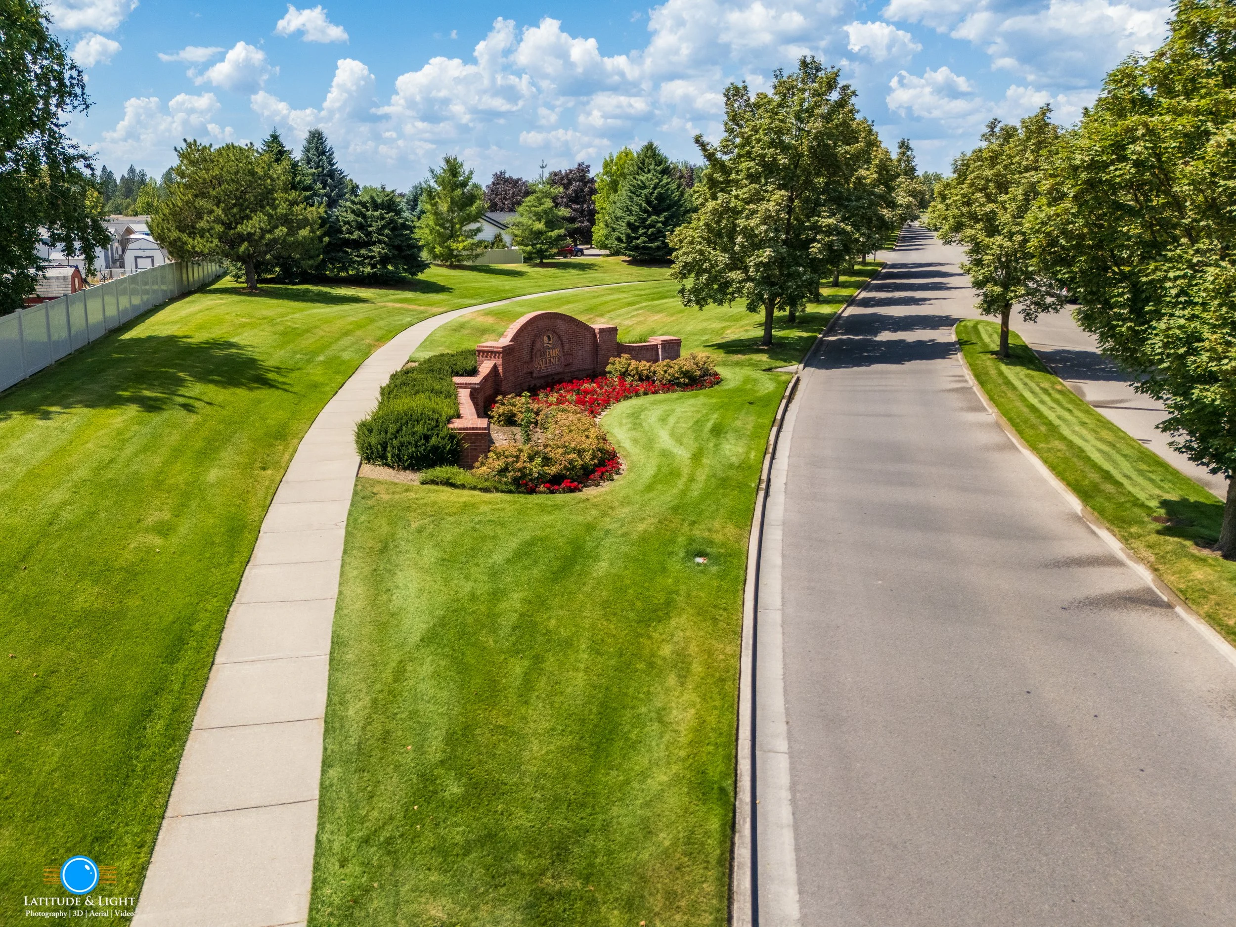 Coeur D'Alene Estates: A suburban neighborhood entrance with a landscaped sign, green grass, trees, and a clean sidewalk on one side of the street.