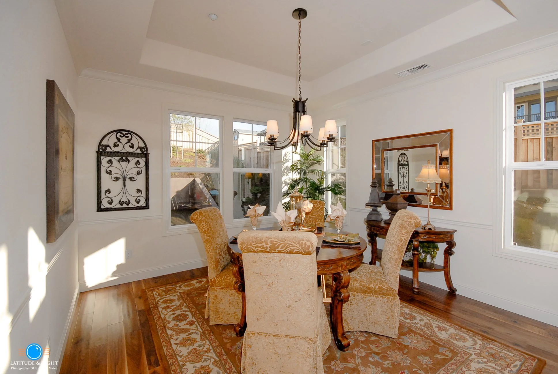 Dining room with a wooden round table, six upholstered chairs, a chandelier, a mirror on the wall, and windows showing an outdoor view.
