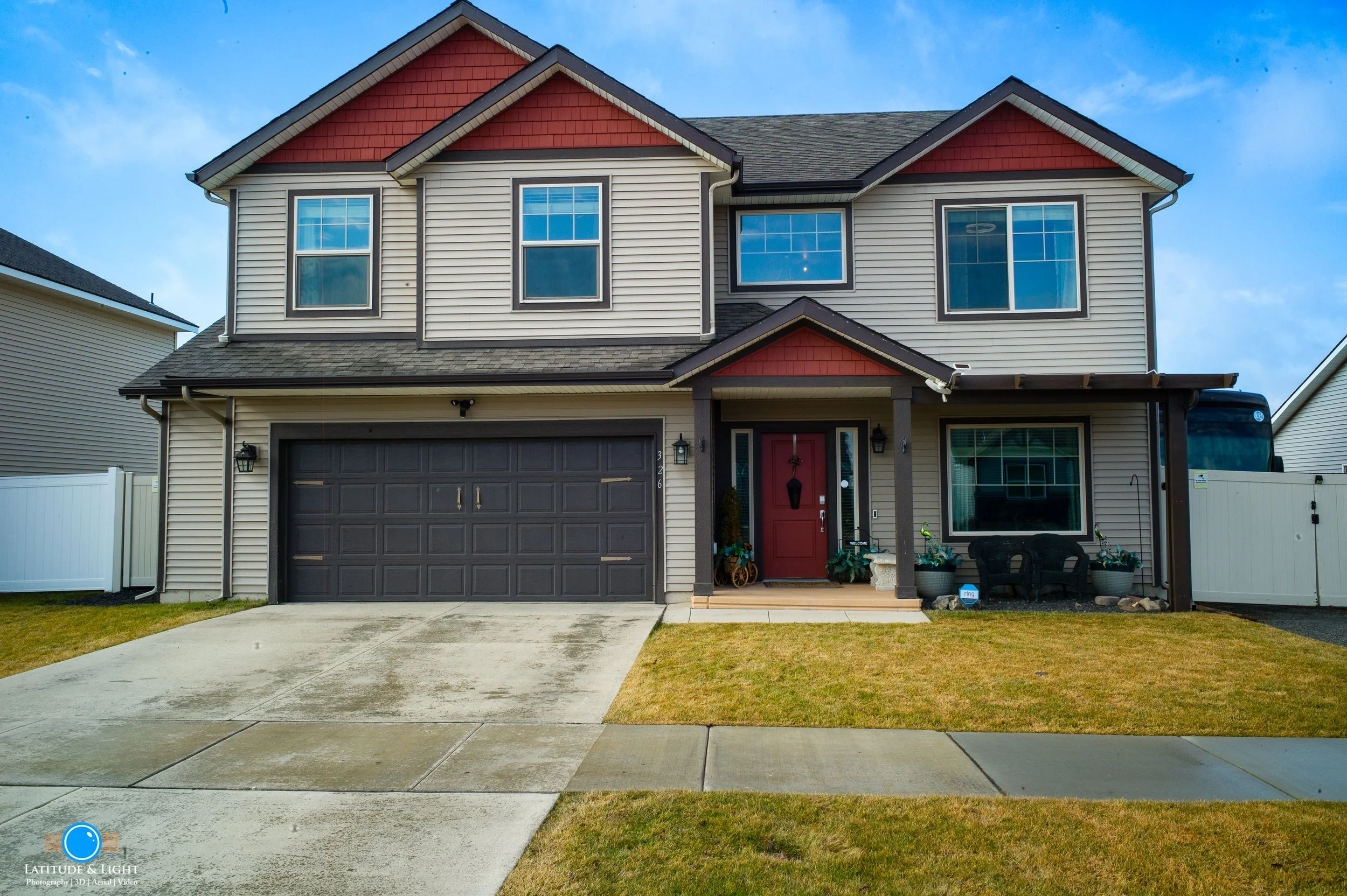 Front view of a two-story house in Airway Heights with beige siding, red accents, a dark garage door, and a red front door, with a grassy lawn and concrete driveway, under a partly cloudy sky.