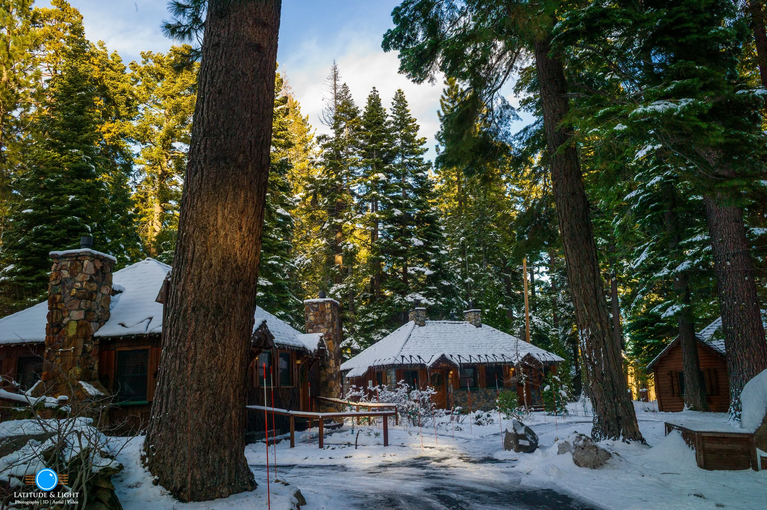 Snow-covered cabins in a Tahoe City resort  surrounded by tall pine trees in a forest setting, with a clear sky and sunlight filtering through the trees.
