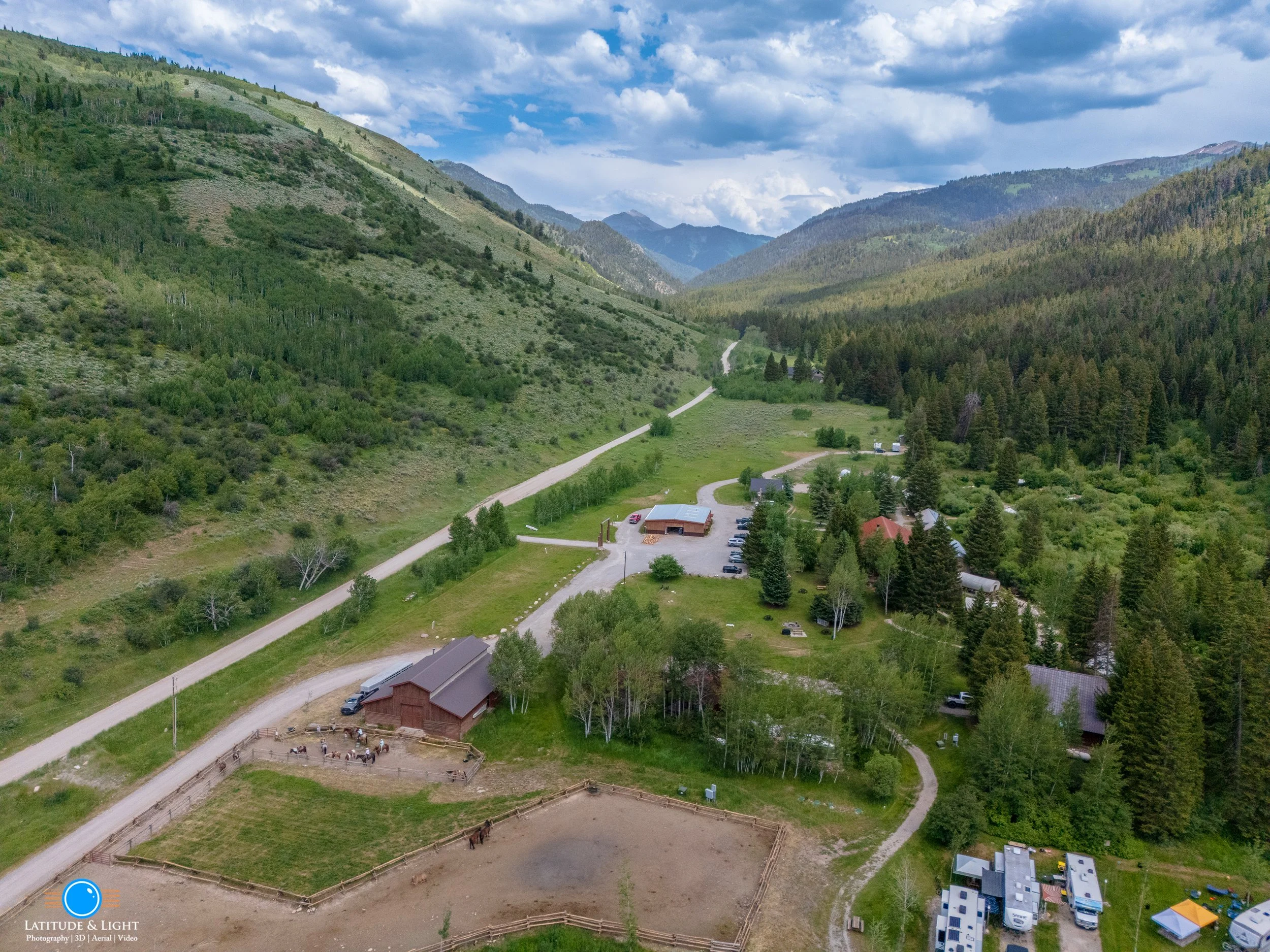 An aerial view of a rural landscape in Victor, Idaho, with green mountains, a dirt road, a barn with a fenced area with horses, and a few buildings among trees.