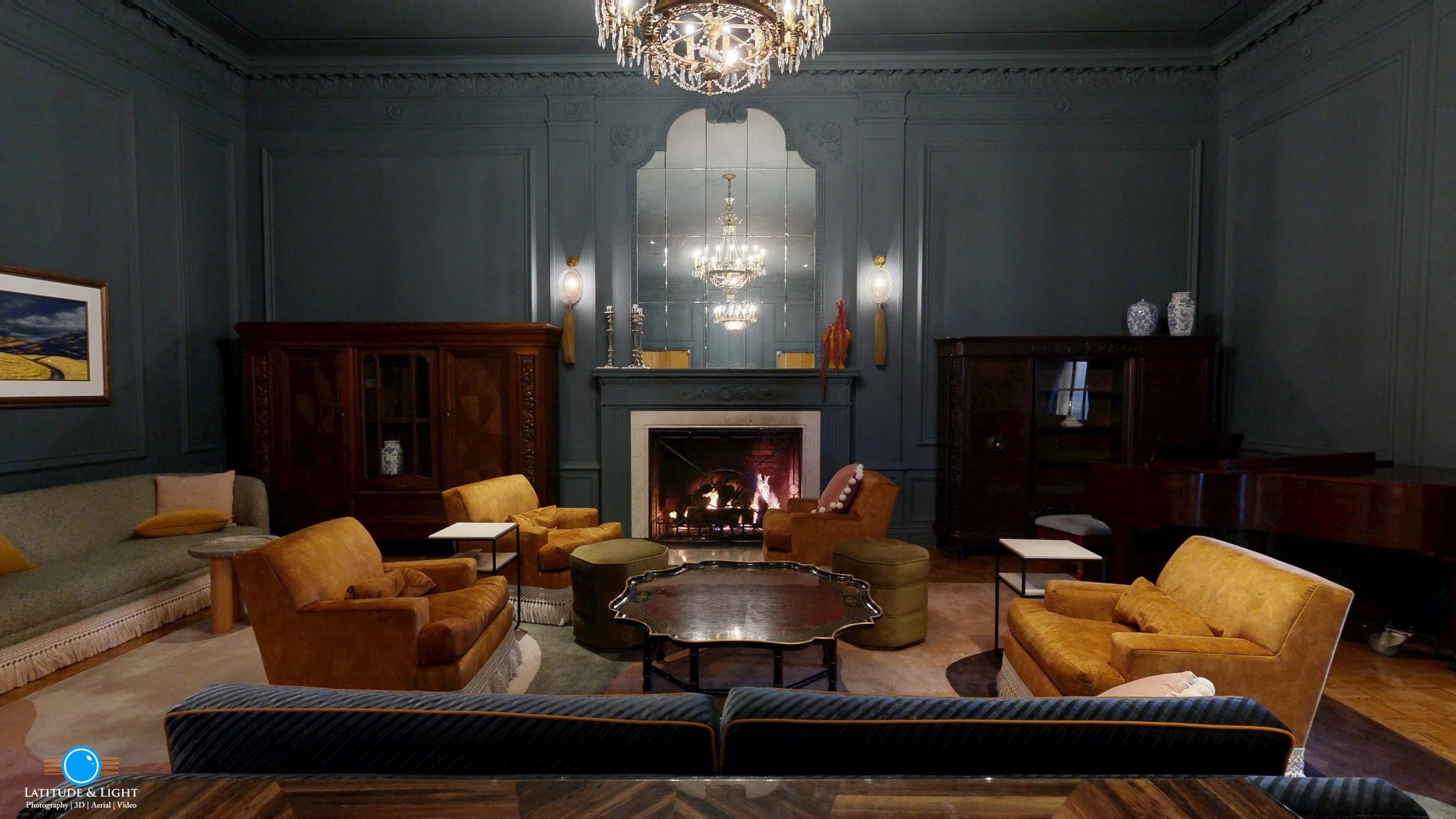 A cozy living room with a lit fireplace, dark green paneled walls, and multiple vintage sofas and armchairs arranged around a wooden coffee table. Decorative items and a chandelier are visible, reflecting in a large mirror above the fireplace.