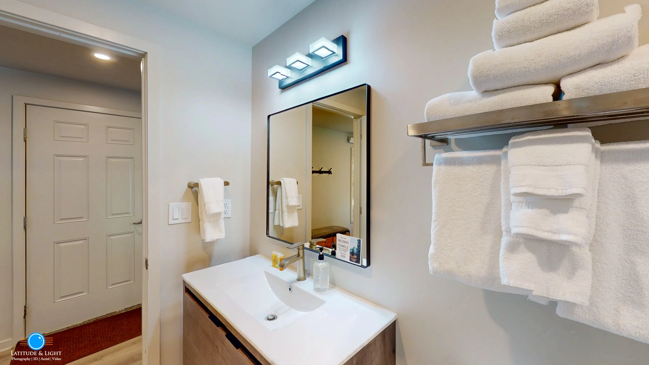 Bathroom with white walls, a rectangular mirror, a modern light fixture above the mirror, a white sink with a wooden cabinet below, a soap dispenser, a small box of toiletries, and shelves with neatly folded white towels. The door to the bathroom is 