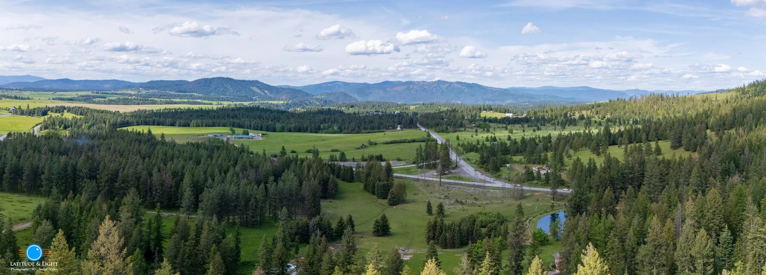 Harrison, Idaho: A panoramic view of a lush green rural landscape with rolling fields, dense forests, a winding road, and distant mountains under a partly cloudy sky.