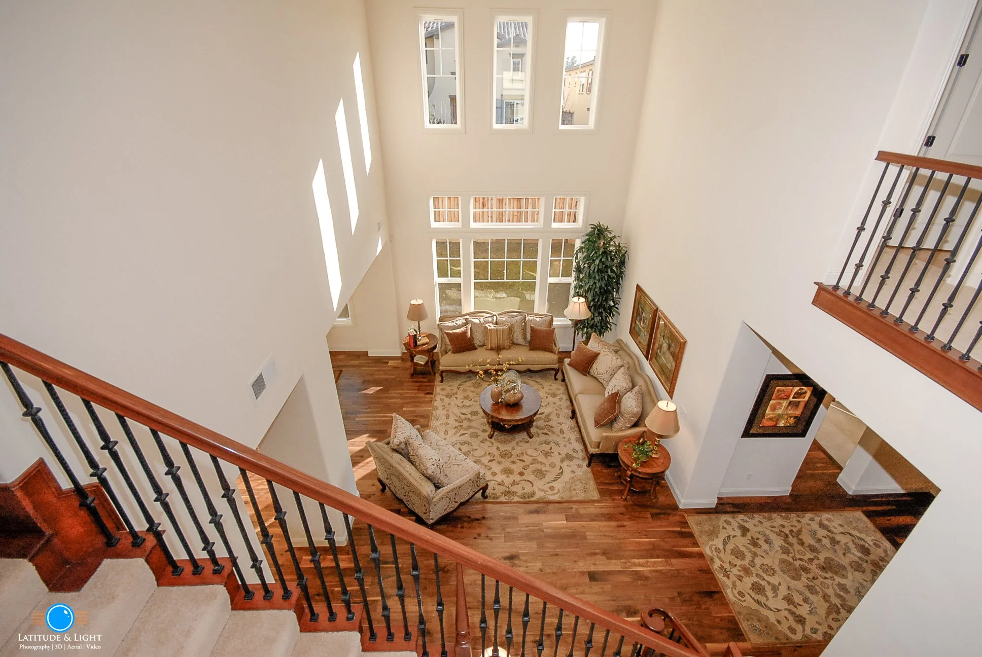 Coeur D'Alene: View from staircase looking down into a two-story living room with high ceilings, large windows, and wooden floors. Furnished with sofas, lamps, and wall art.