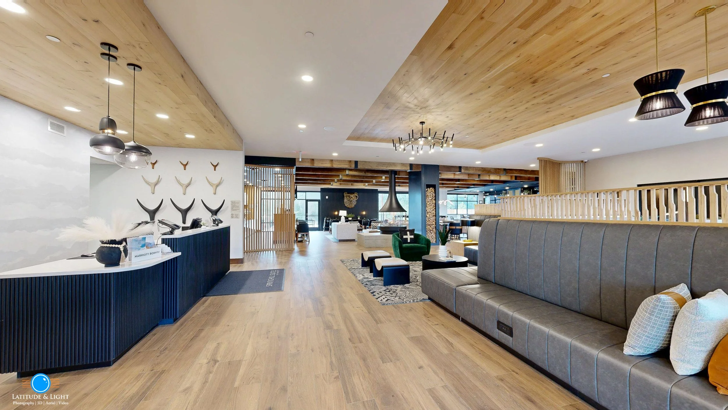 Interior of a modern hotel lobby in Sandpoin with a reception desk on the left, decorated with antler wall art and tables with umbrellas, a comfortable seating area with green and gray chairs, a fireplace, and large windows providing natural light.