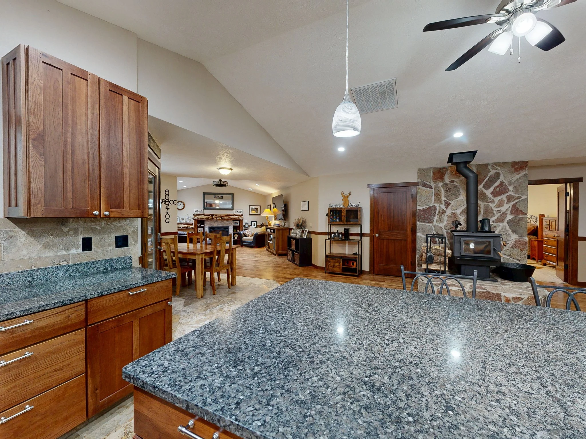 Open concept kitchen and living room with granite countertops, wooden cabinets, a stone fireplace, and a wood stove with a chimney.