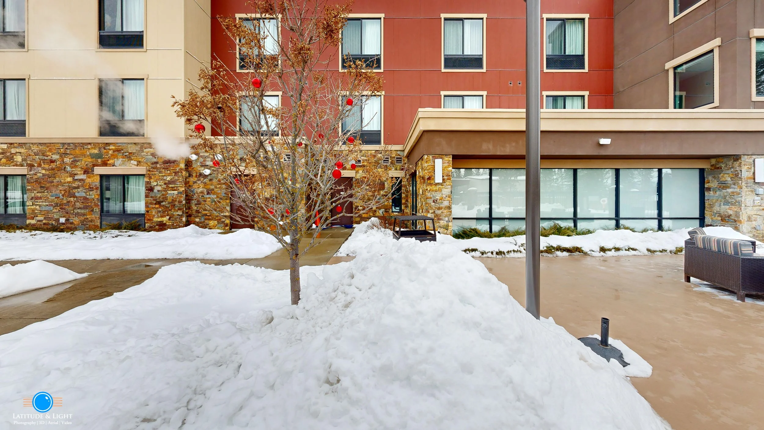 The front of a Kalispell, Montana ski hotel with a stone and stucco exterior, snow on the ground, a leafless tree with red ornaments, a lamppost, and outdoor seating.