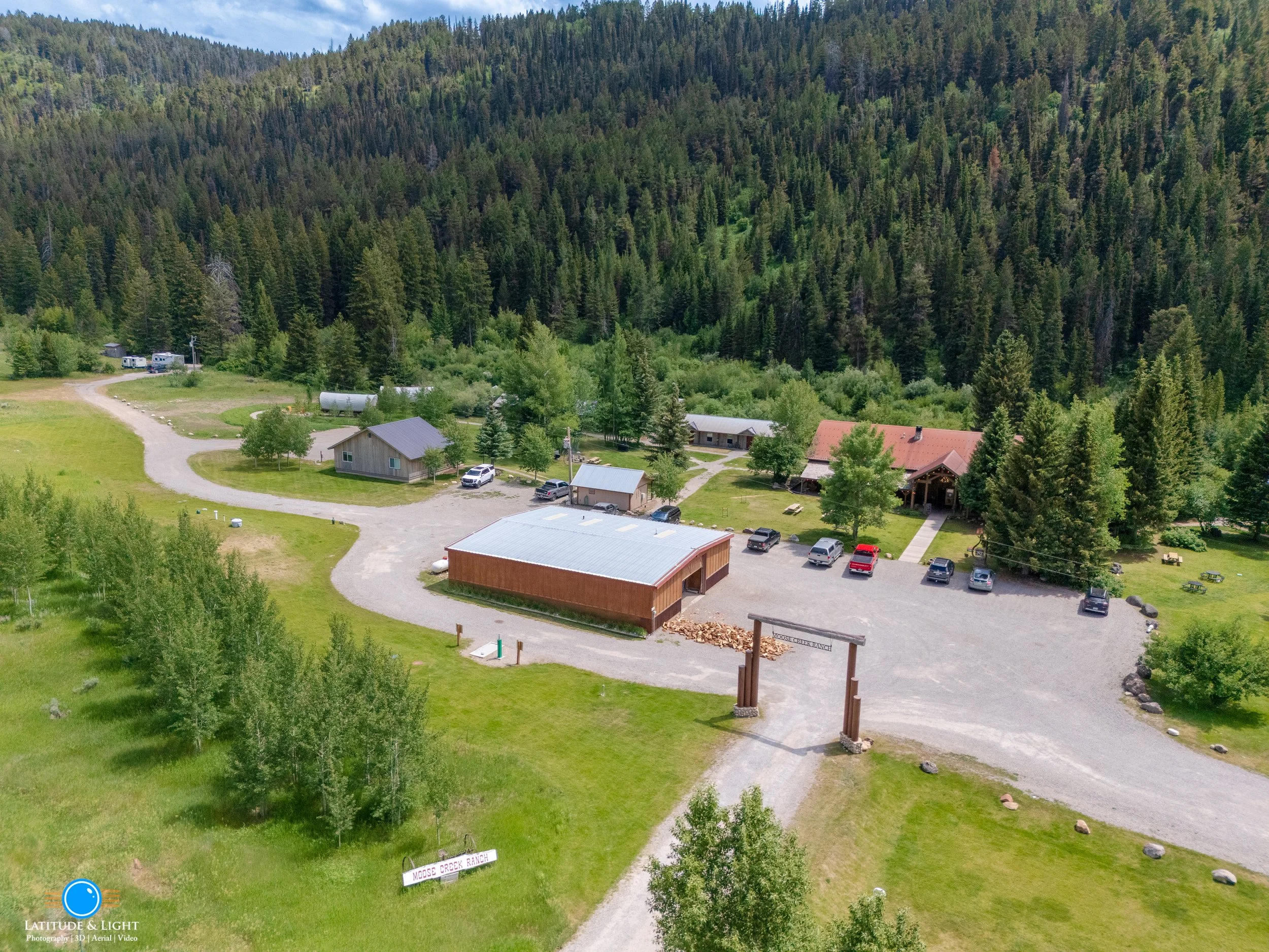 Aerial view of Moose Creek Ranch in Victor, Idaho, featuring a gravel driveway, a wooden building, several parked cars, and a lush green landscape surrounded by dense forest.
