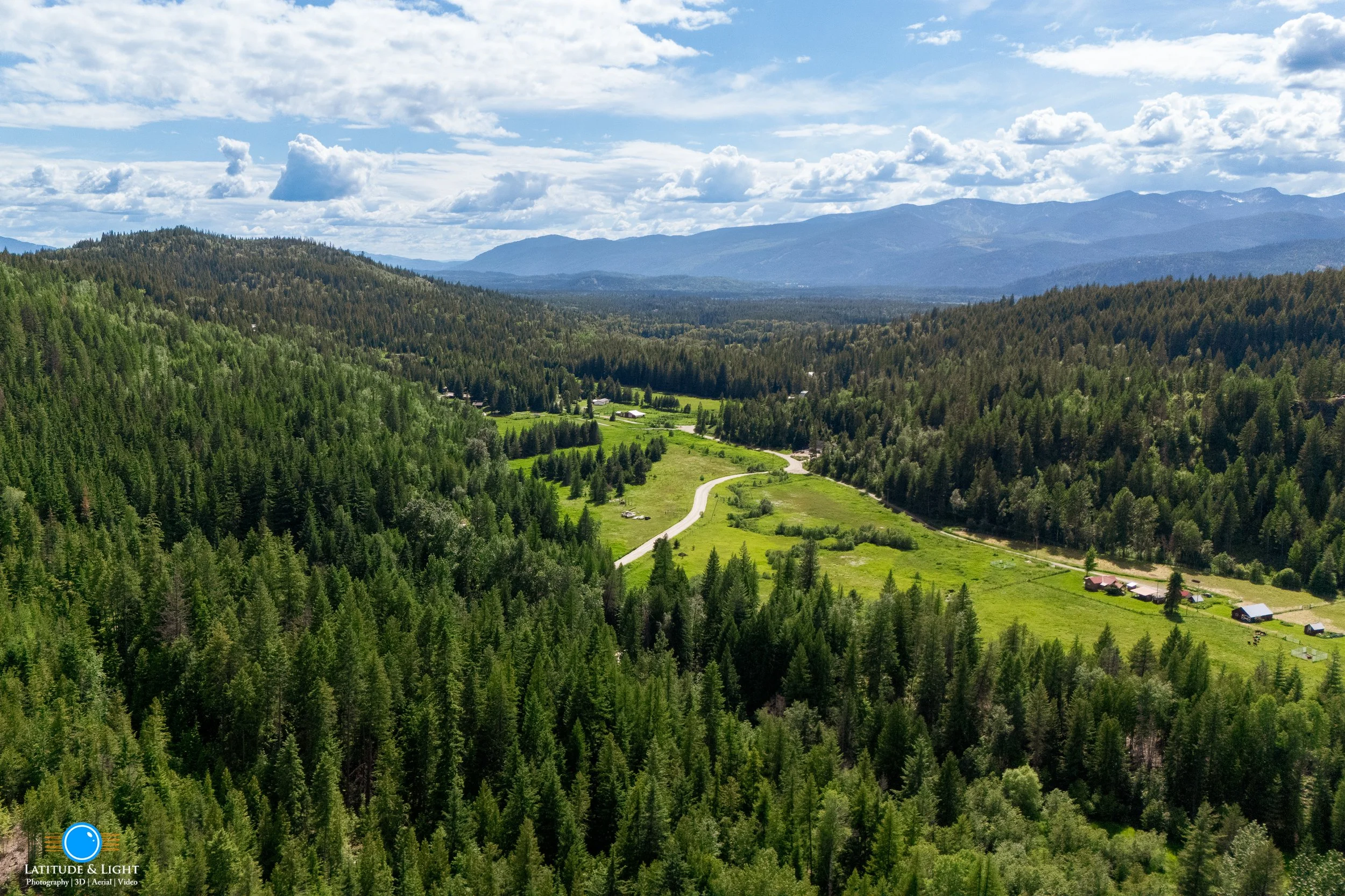 Harrison, Idaho: A scenic aerial view of a lush green valley with a winding road, surrounded by dense forests and distant mountains under a partly cloudy sky.