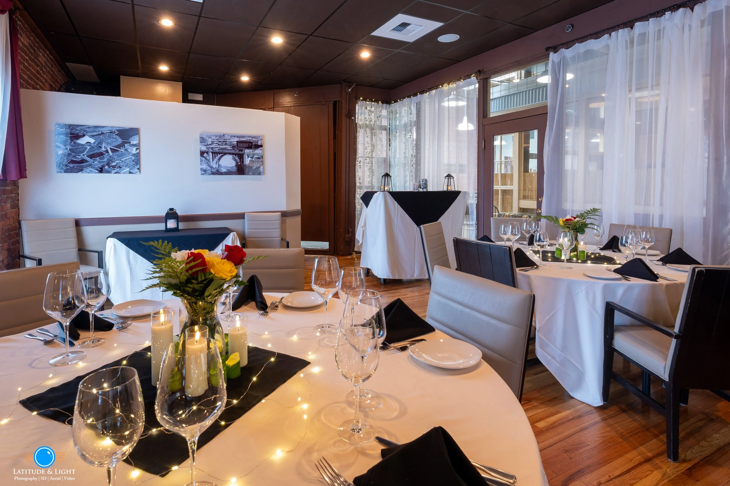 A decorated private dining area in a Spokane restaurant  with round tables set with white tablecloths, black napkins, wine glasses, plates, and silverware, featuring floral centerpieces and candles, windows with sheer curtains allowing natural light.