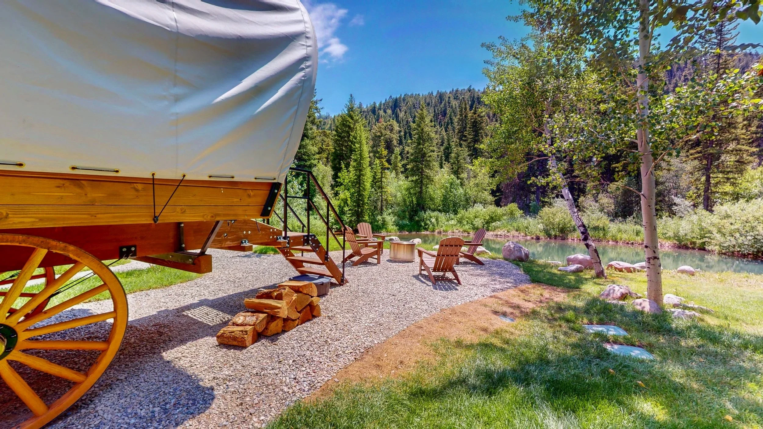 A rustic outdoor scene in Montana with a covered wagon, Adirondack chairs, and a small fire pit beside a river, surrounded by trees and mountains on a sunny day.