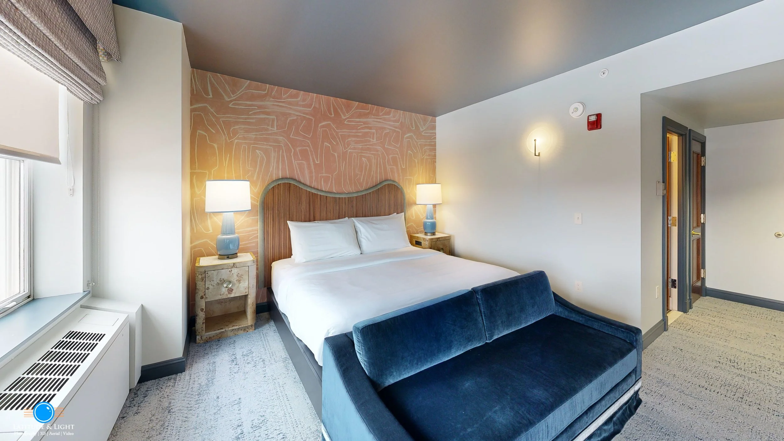 A bedroom with a bed, two nightstands with lamps, a blue velvet bench at the foot of the bed, a window with blinds, and an open doorway leading to another room.