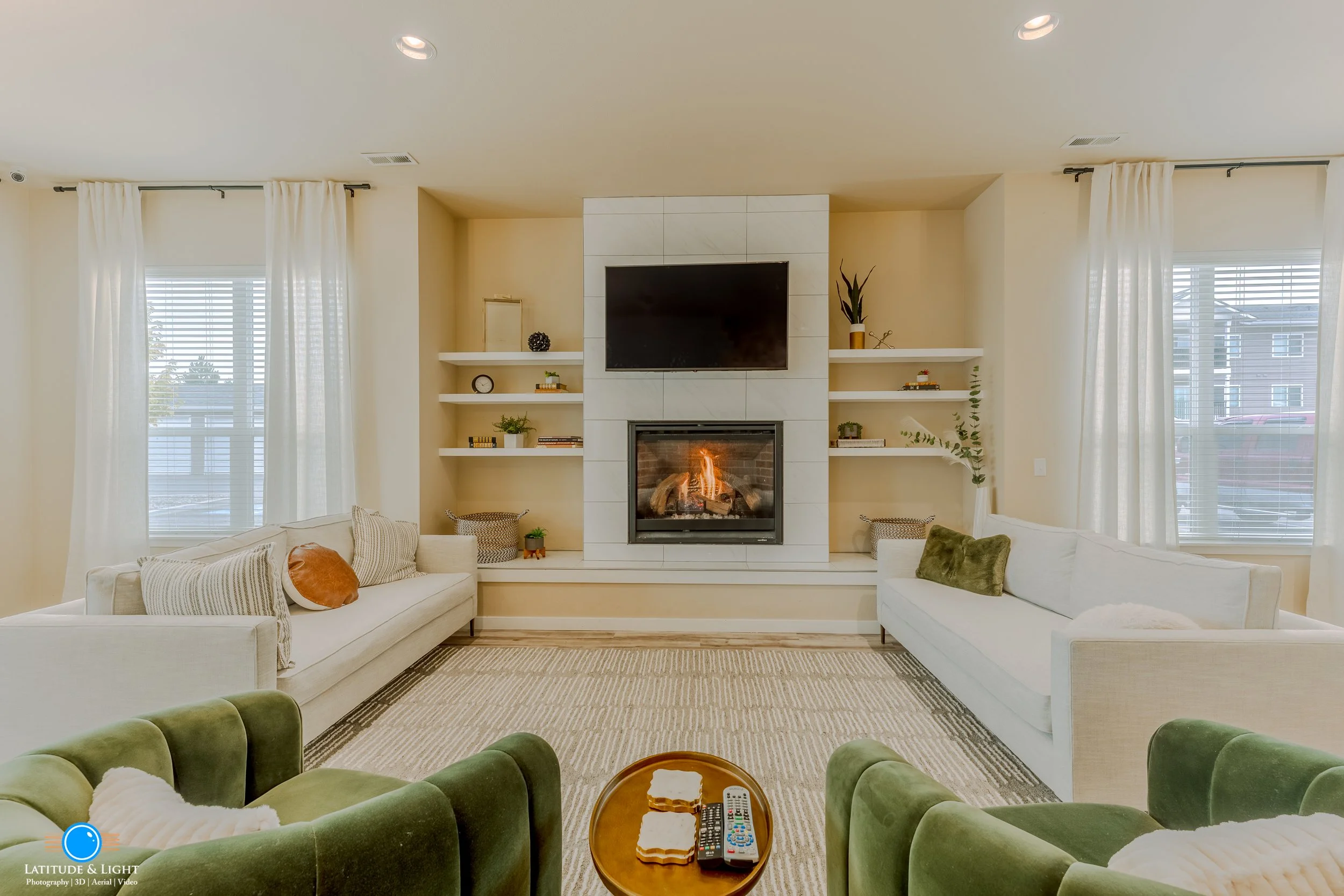 Community center in a Post Falls condo development with two white sofas, green velvet armchairs, a fireplace with a television above it, and shelves with decorative items and plants, illuminated by natural light through large windows.