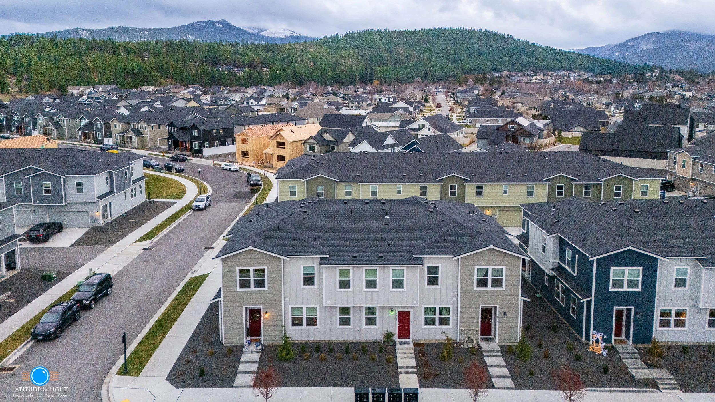 Aerial view of a suburban neighborhood with new houses, some under construction, surrounded by green hills and mountains in the background.