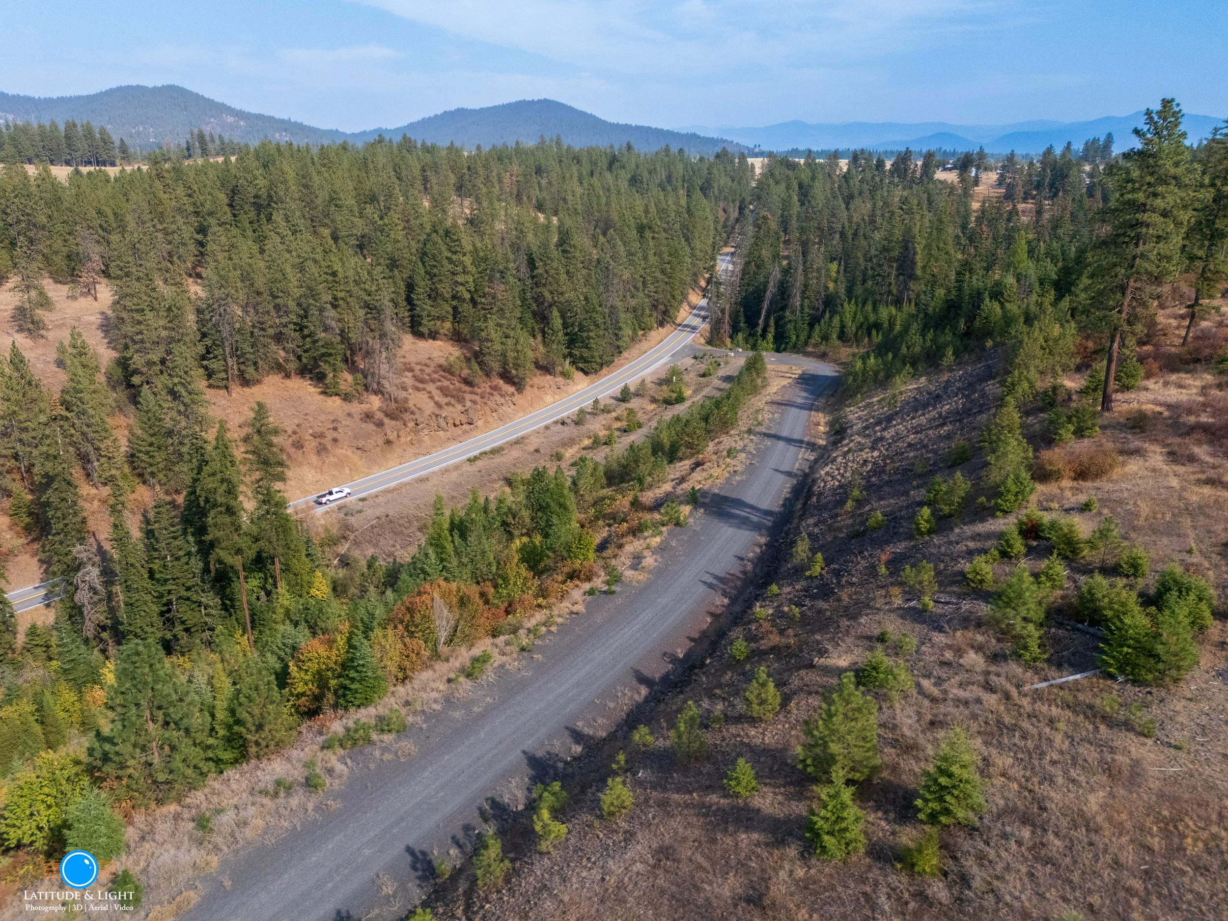 Harrison, Idaho: An aerial view of a mountainous landscape with a winding road and a small dirt road running parallel. The scene includes dense evergreen trees on both sides of the roads and distant mountains in the background under a blue sky.