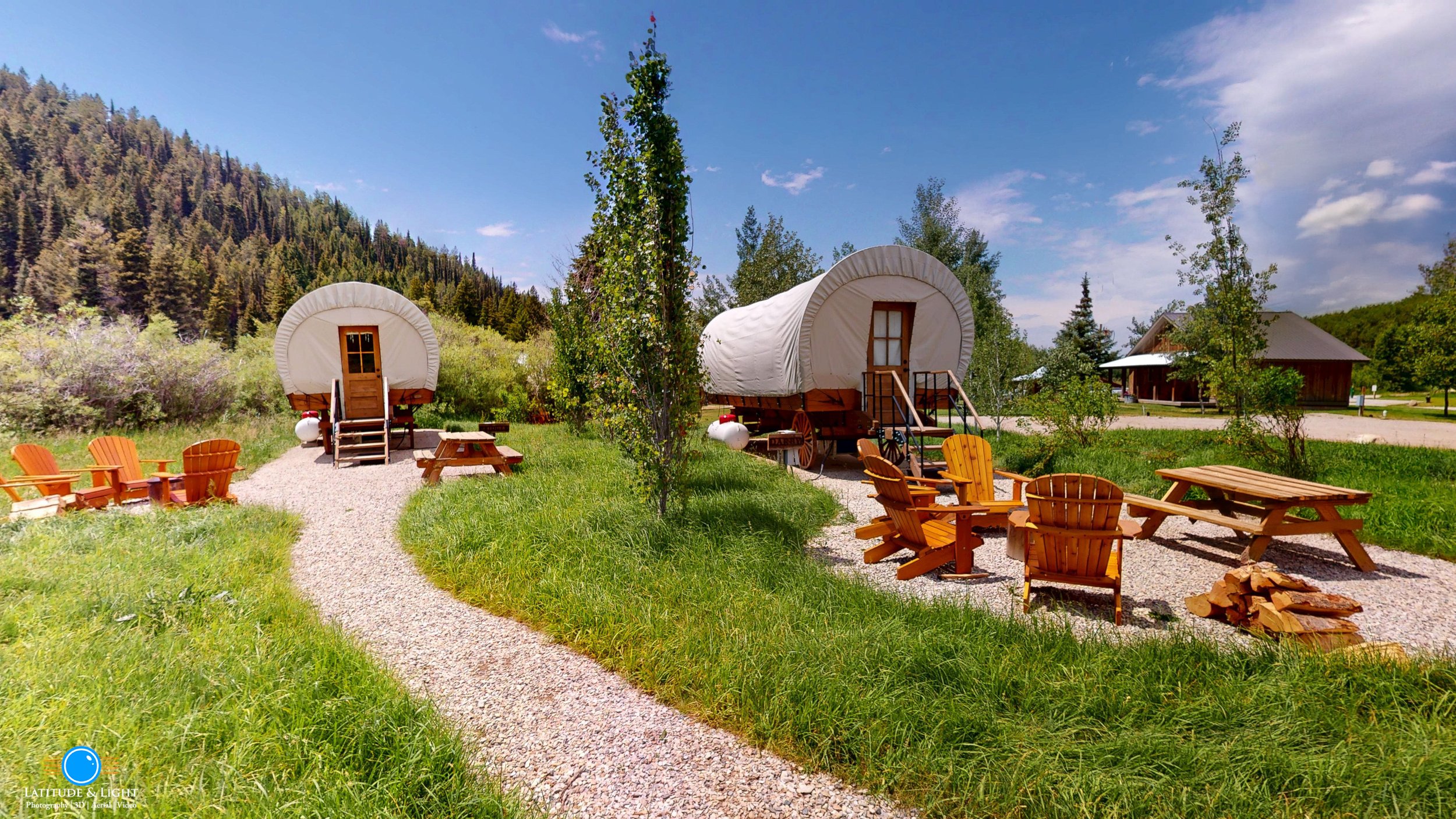 Scenic outdoor area in Victor, Idaho with two white covered wagons, surrounded by green grass, trees, and mountains in the background. Wooden Adirondack chairs, picnic tables, and a firepit are arranged around a gravel pathway on a sunny day.
