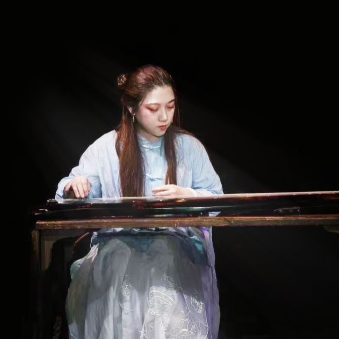 Clover Hu with long brown hair styled in a traditional updo, playing a traditional Chinese guqin instrument on a dark stage, wearing a light blue traditional Chinese dress.