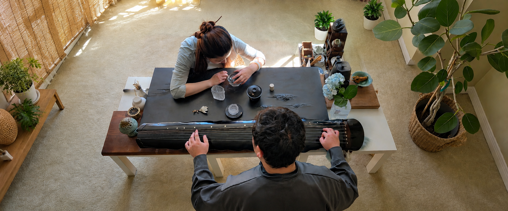 A woman and a man are sitting at a table in a room filled with plants. The woman is working on a black piece of art or craft, and the man is playing a qin.